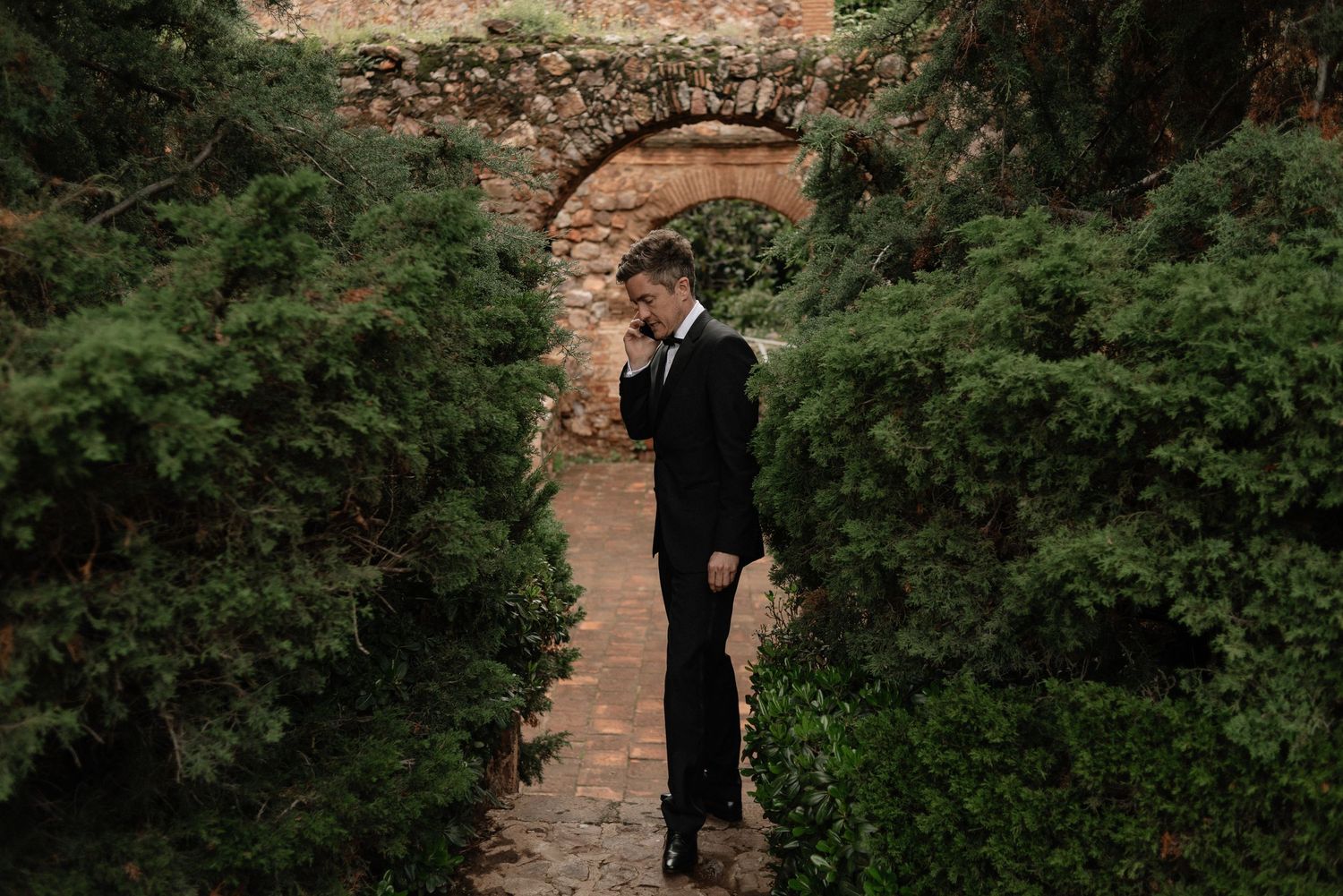 A silhouetted figure stands beneath a rustic stone archway surrounded by lush green foliage.