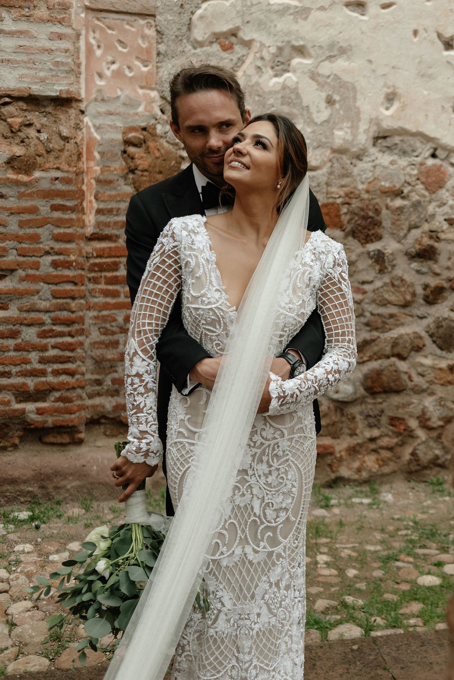 A romantic moment between newlyweds features an elegant lace wedding dress and flowing veil against a rustic stone wall.