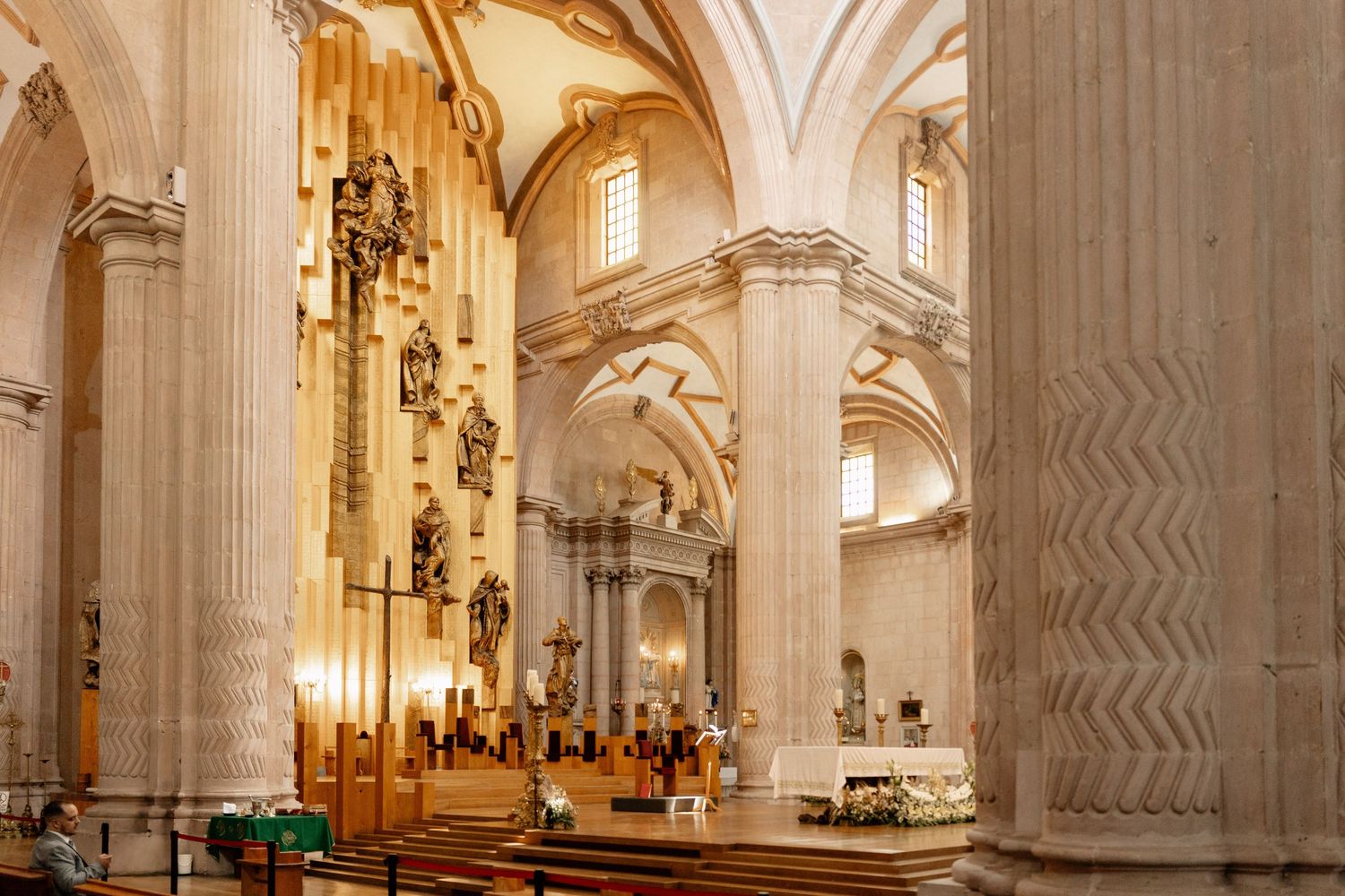 Interior view of an ornate cathedral with white stone columns, vaulted ceilings, and golden chandeliers illuminating the space.