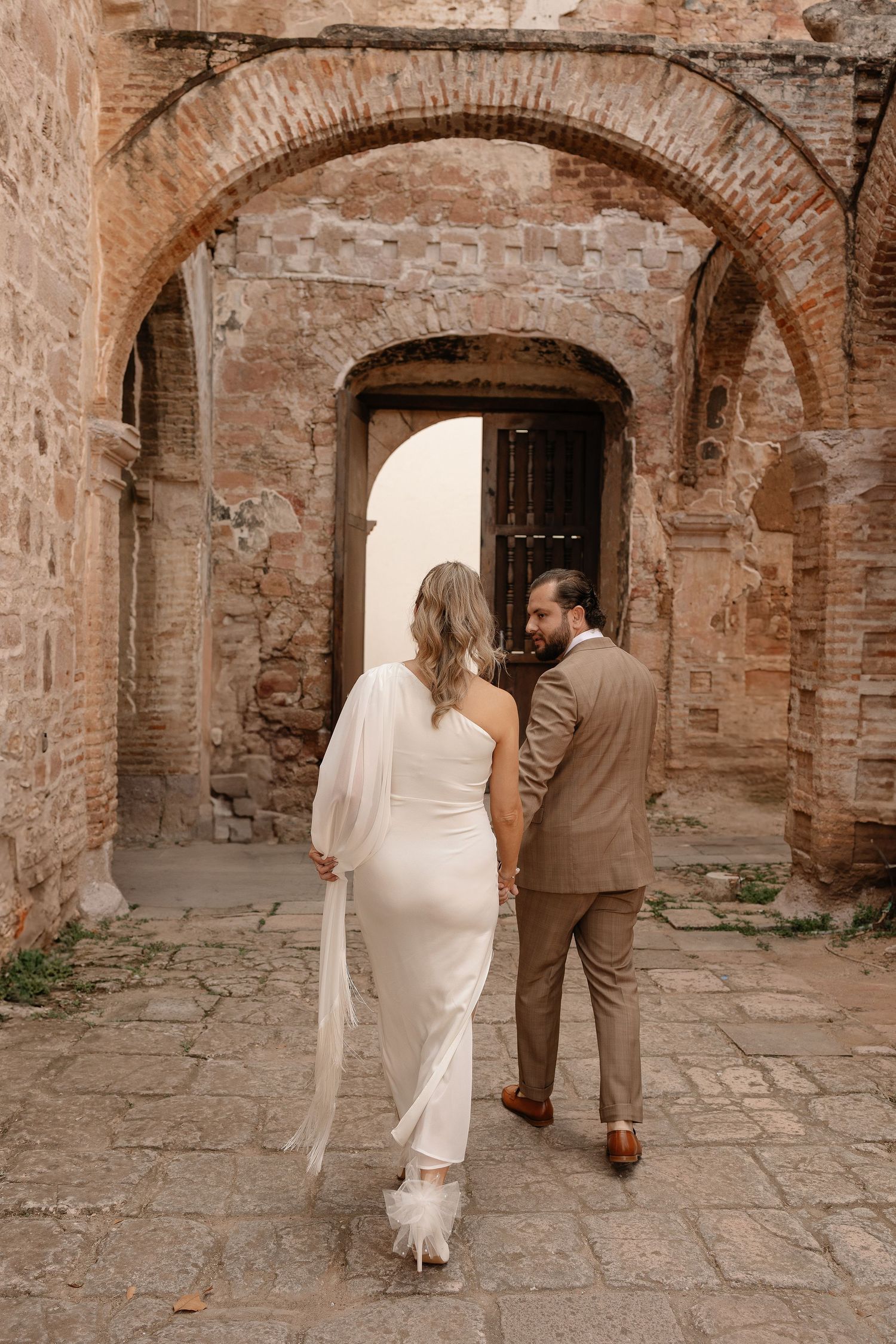 A couple in wedding attire walks through a rustic stone archway corridor in a historic building.