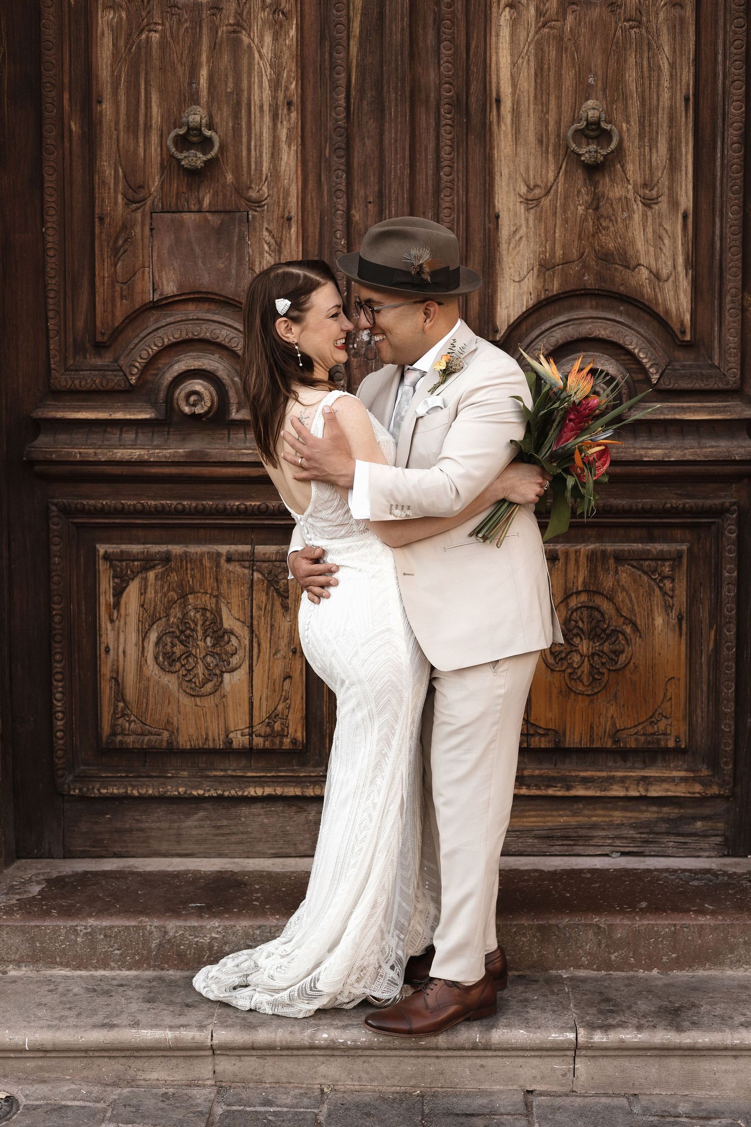 A couple embraces against ornate wooden cathedral doors in wedding attire.