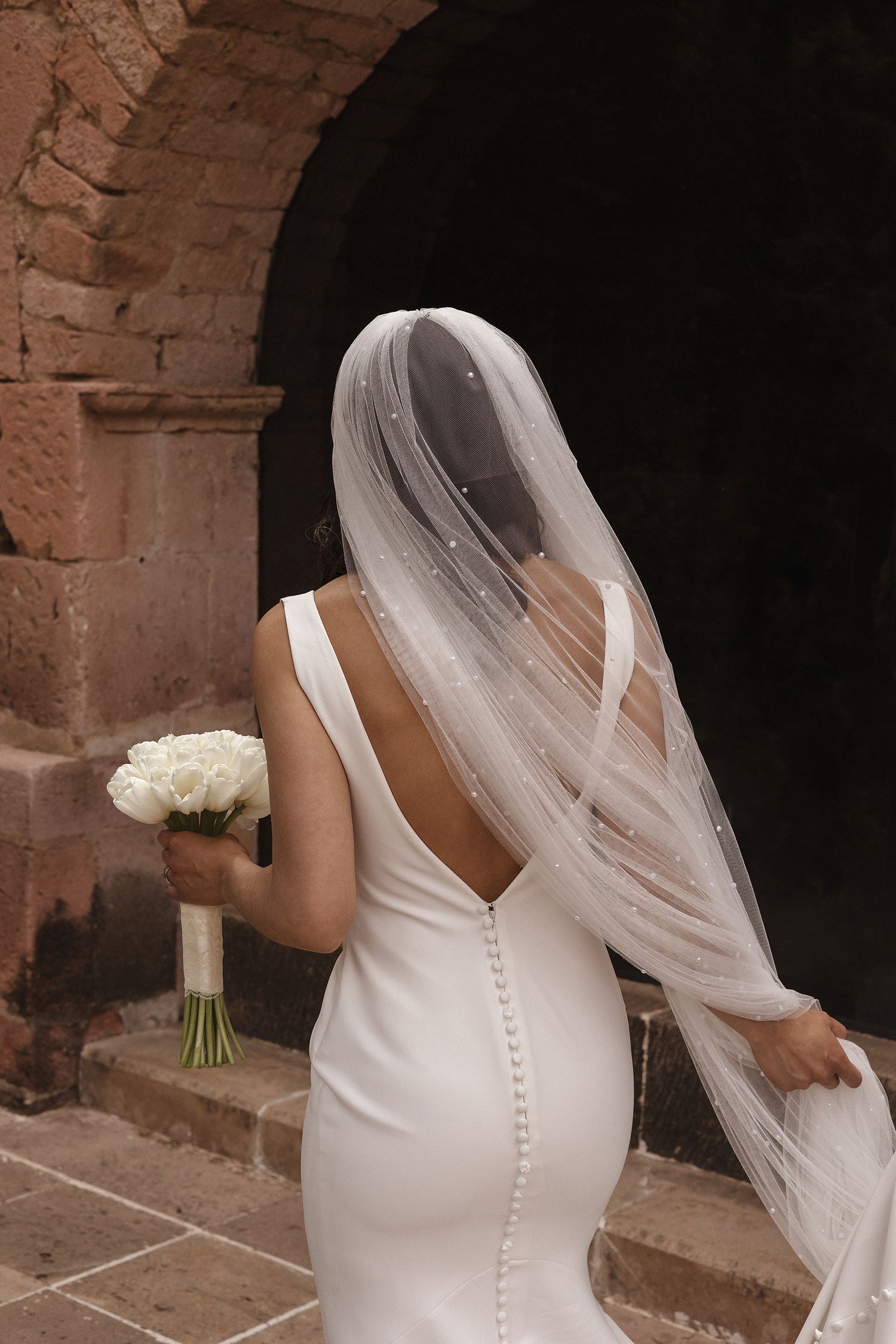 Elegant white wedding dress with button details and flowing veil shown from behind in a rustic brick setting.
