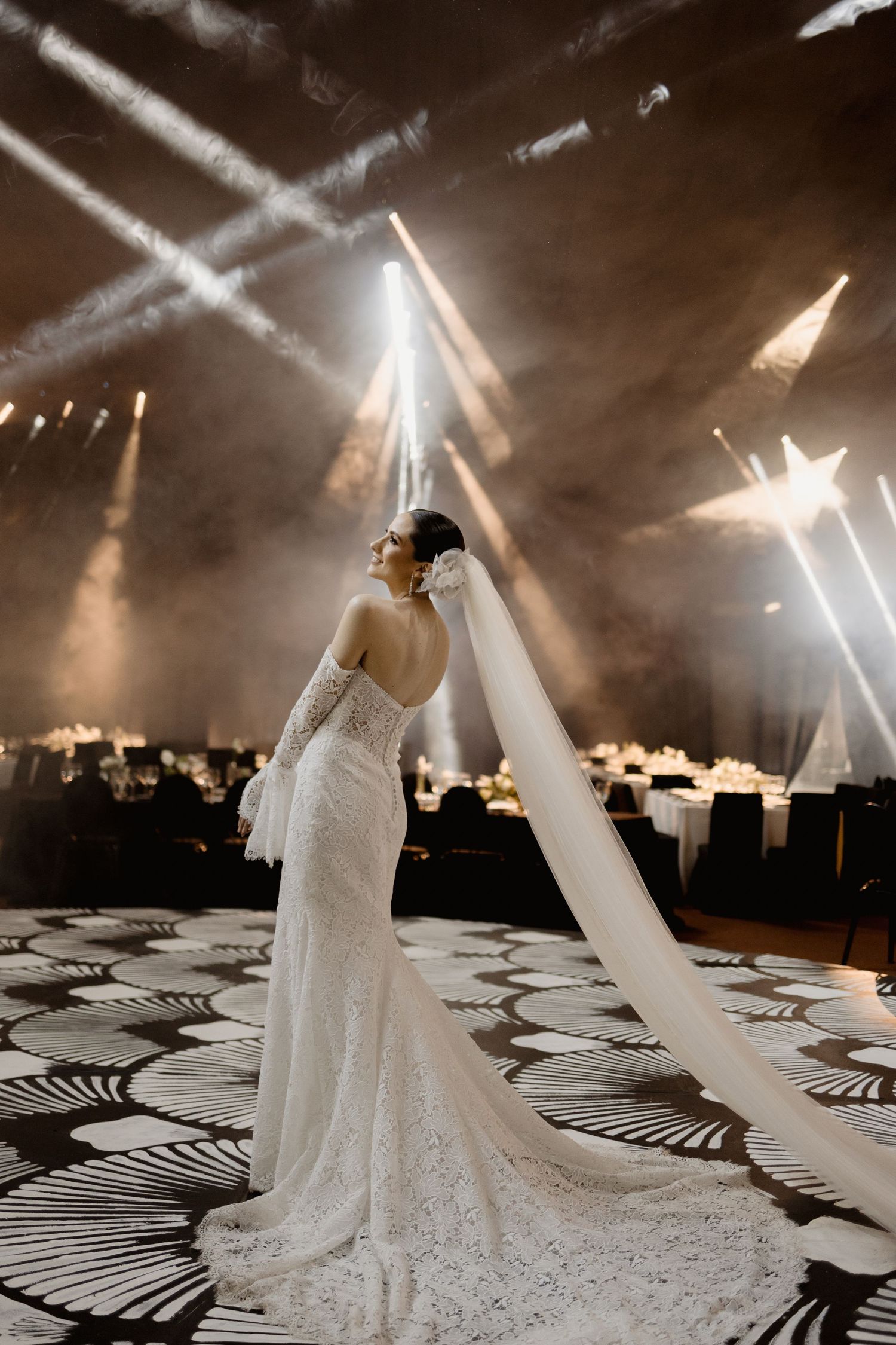 A dramatic wedding scene with spotlights illuminating a white lace gown against a black and white geometric floor pattern.