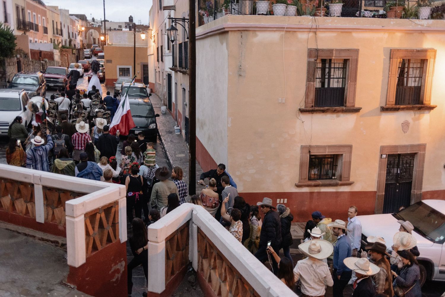 People gather on a street corner in a colonial Mexican town with peach-colored buildings and Spanish architecture.