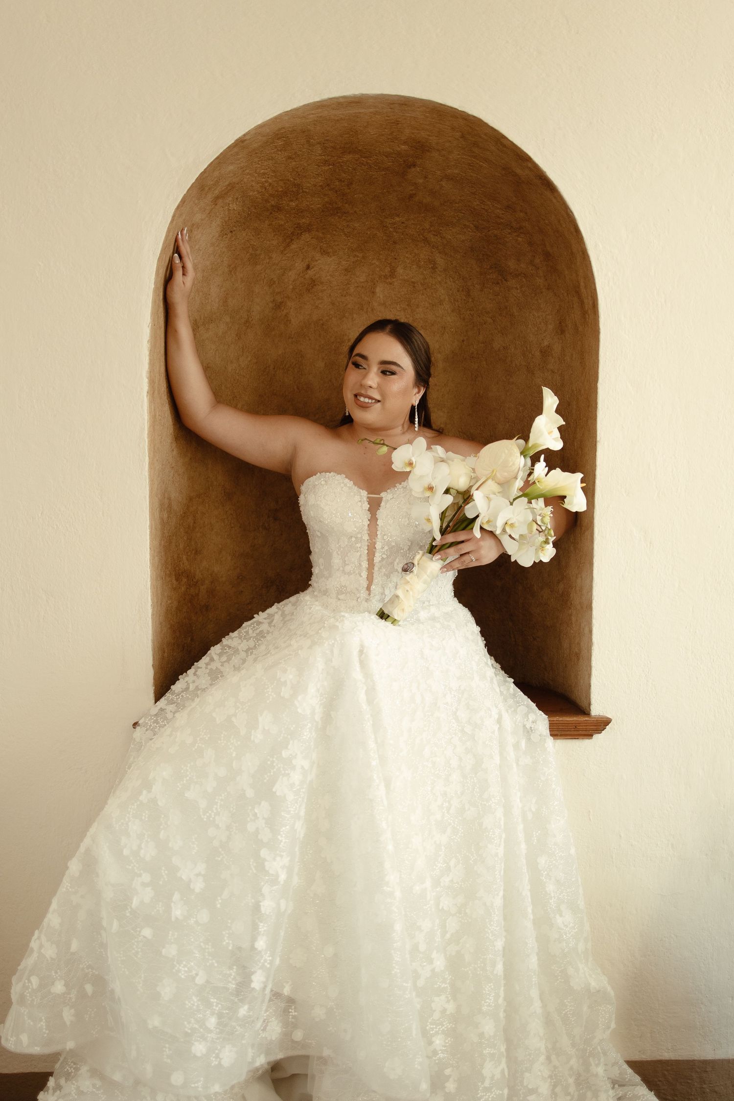 A stunning white ball gown wedding dress with floral details shown in multiple poses against a brown alcove backdrop.