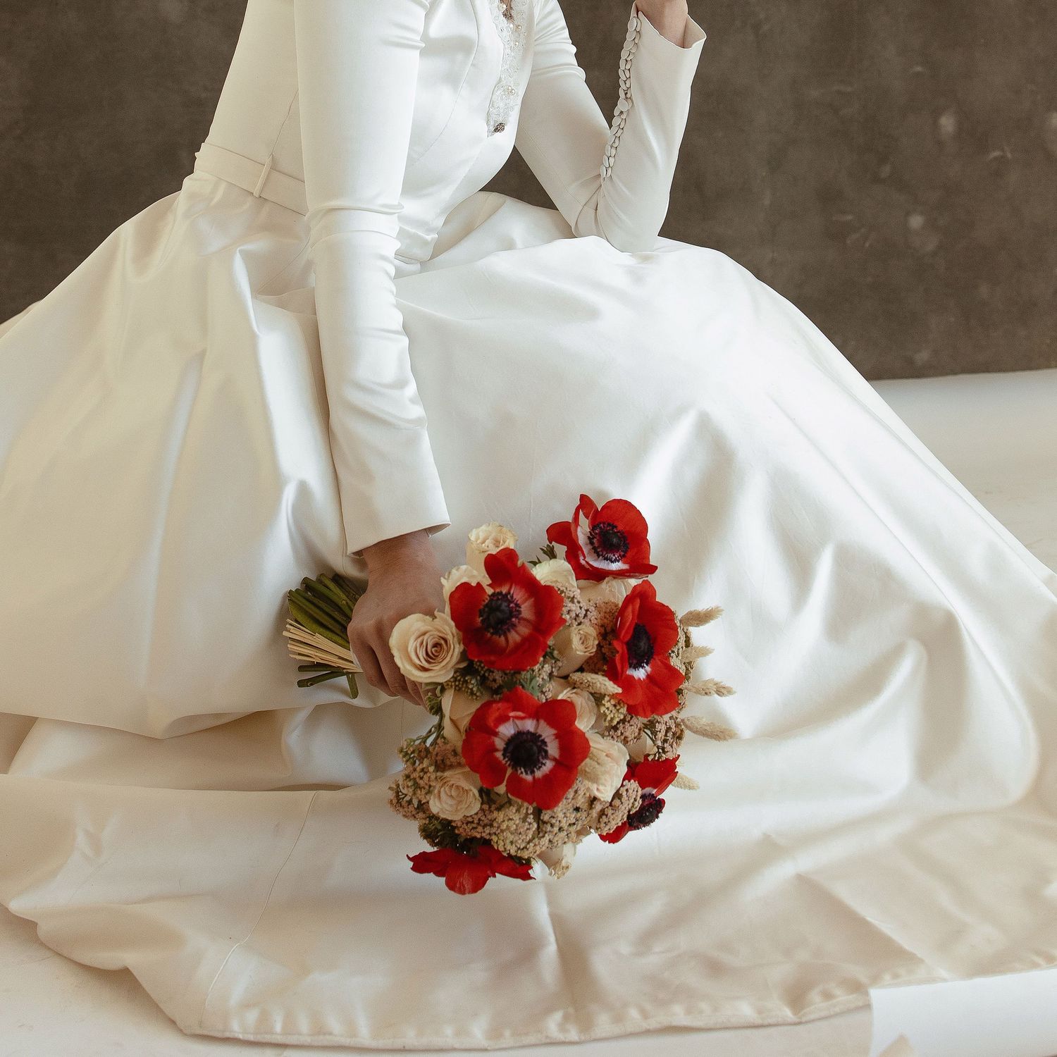 A cascading bridal bouquet with vibrant red anemone flowers and blush roses against a white wedding dress background.