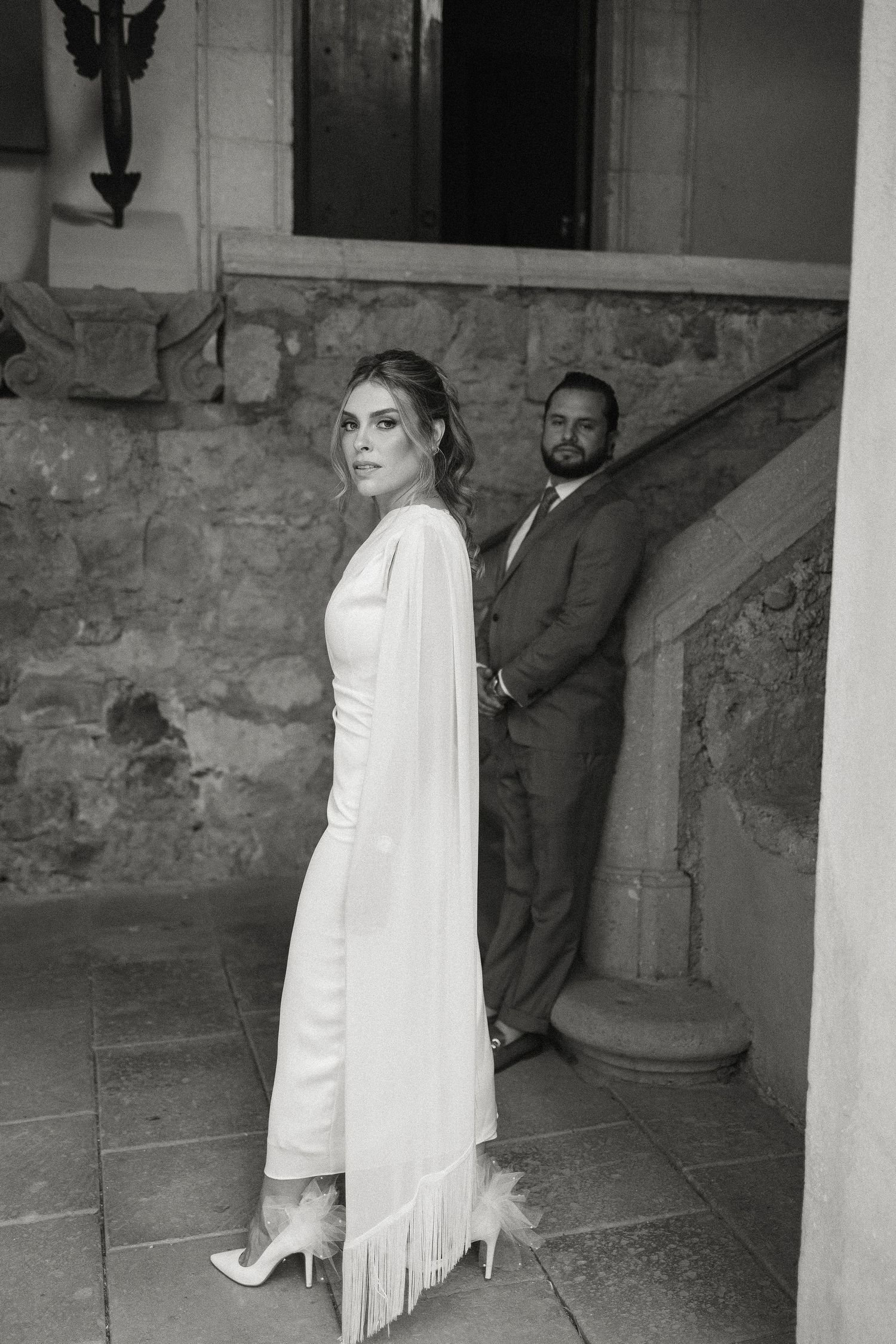 Black and white wedding photo showing couple in elegant attire posing on stone staircase with vintage architecture.
