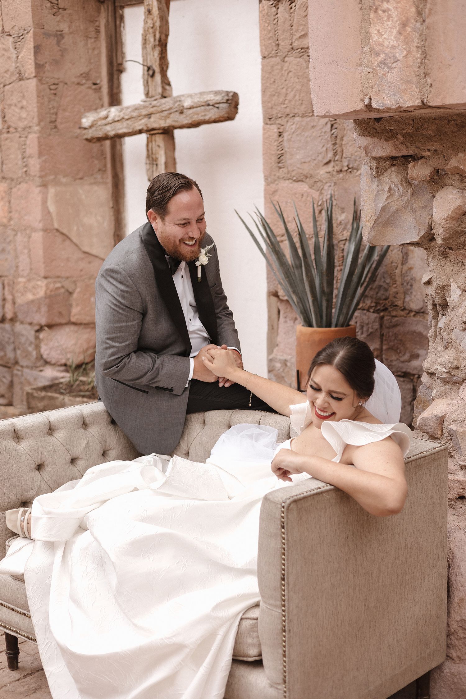 A joyful wedding couple shares a playful moment on a beige couch in a rustic stone setting.