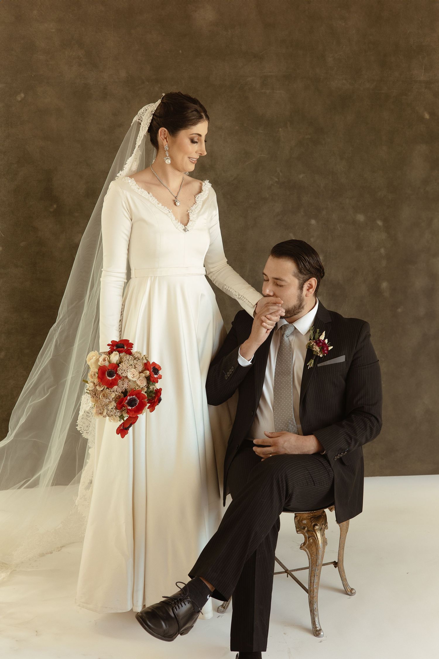 Elegant wedding portrait with bride in long-sleeved white gown holding red bouquet and groom seated in dark suit.