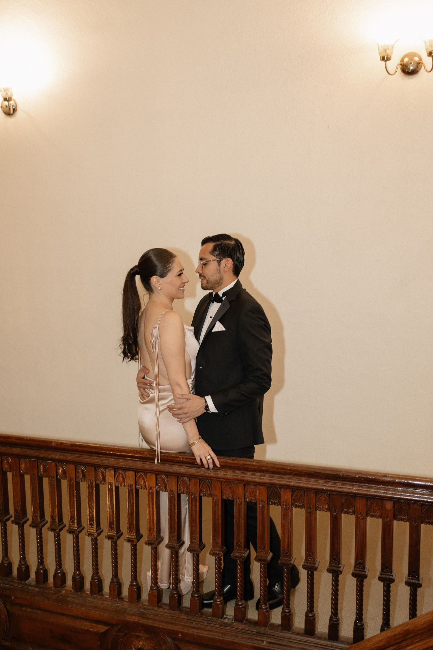 A romantic couple shares a tender kiss on a staircase landing with wooden railings and warm lighting.