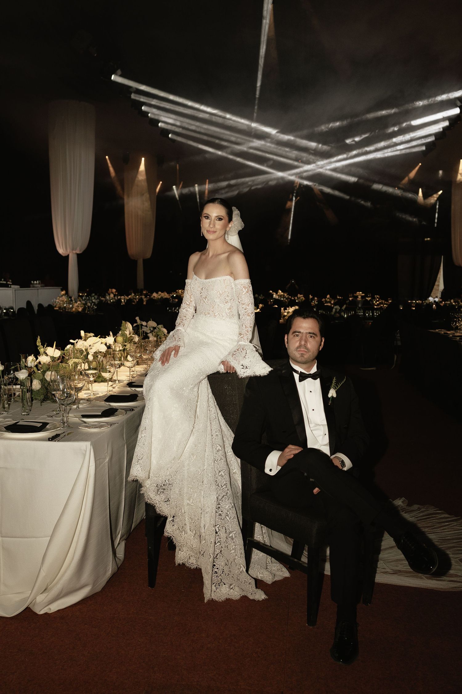 Elegant wedding reception table setting with dramatic lighting and white decor under crossed beams at night.