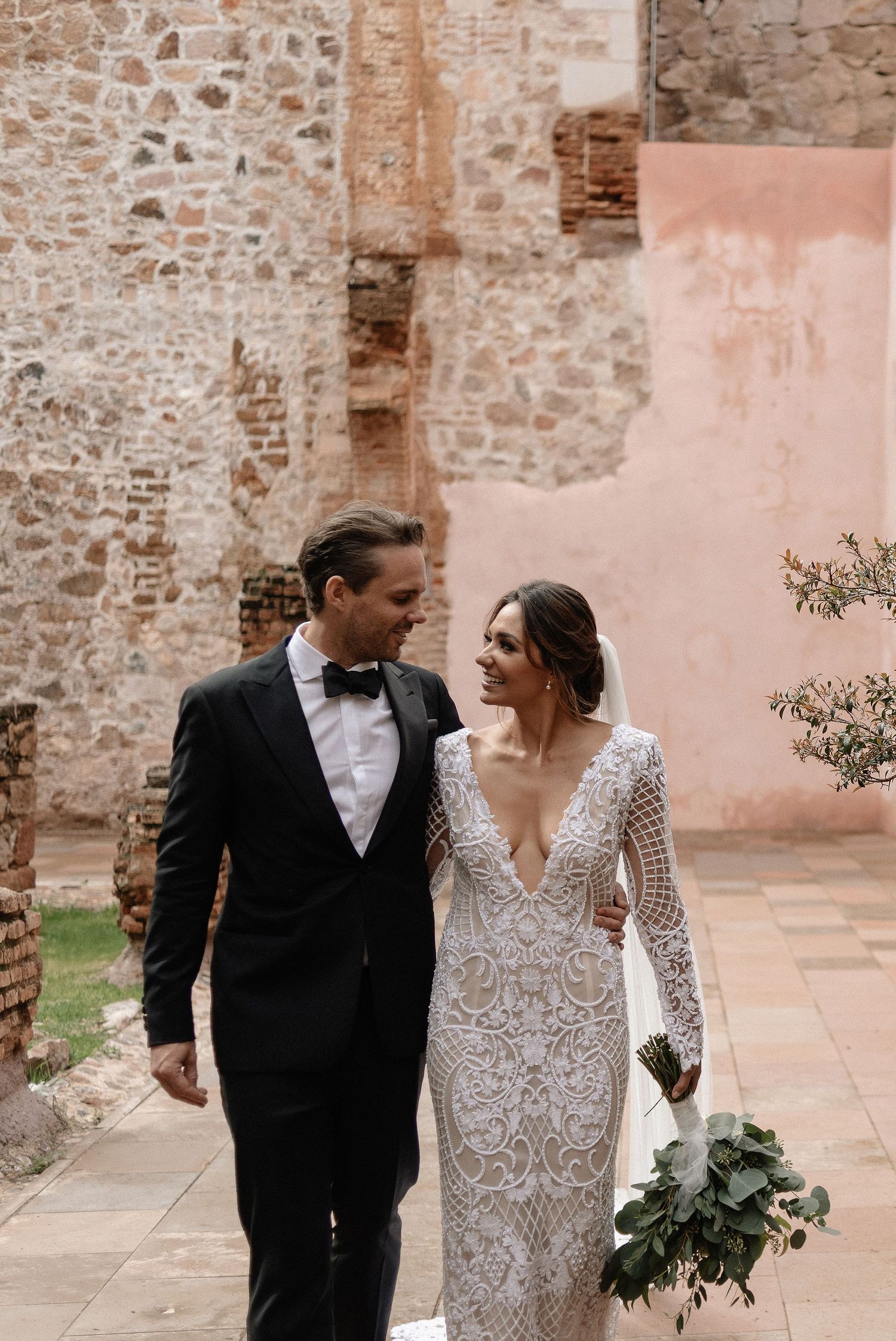 A couple in formal wedding attire walk together along a stone path beside a textured pink and beige wall.
