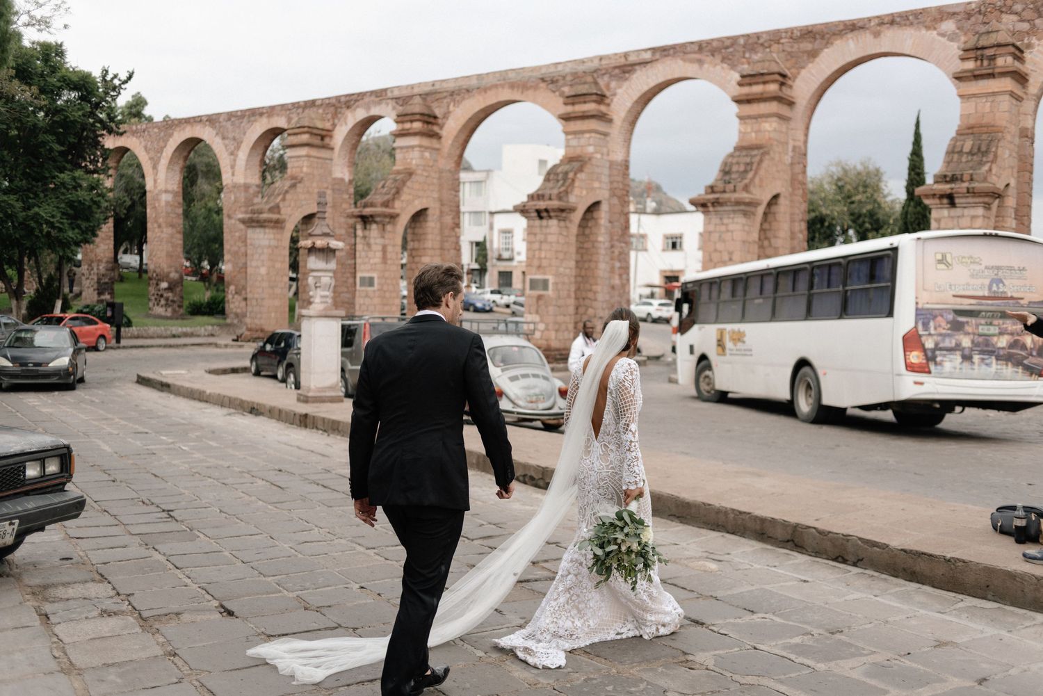 A couple in wedding attire walks along a historic stone aqueduct in Morelia, Mexico with a tour bus parked nearby.