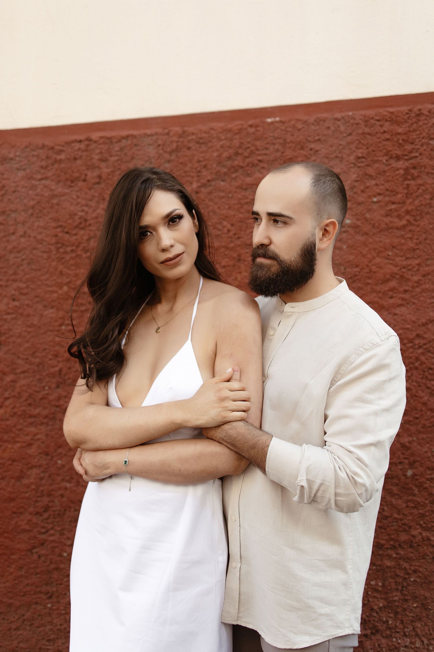 A couple poses romantically against a rustic red wall while wearing coordinating white outfits.