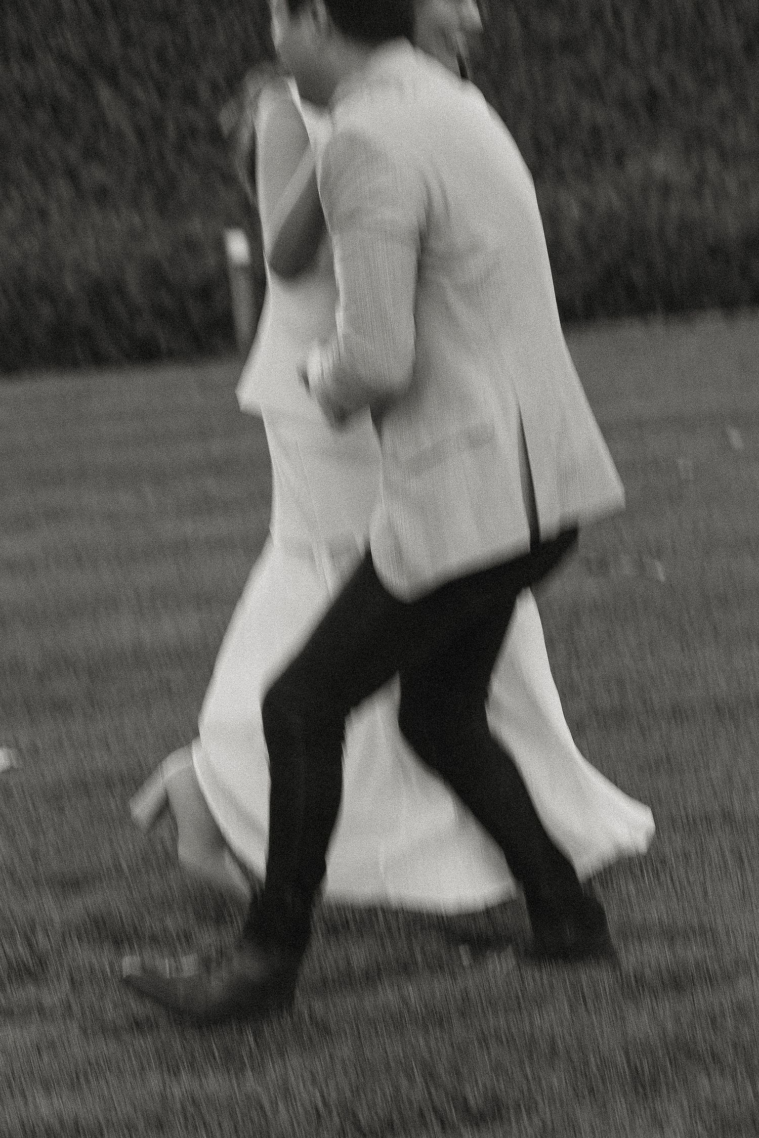 A black and white motion-blurred photograph of two people dancing together on grass outdoors.