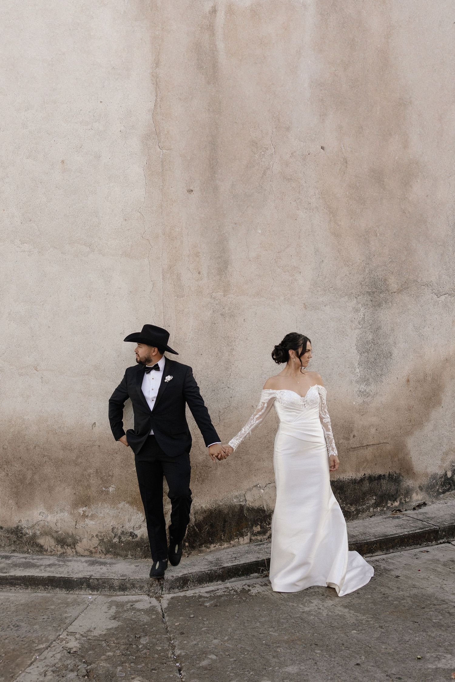 A couple in wedding attire holds hands while walking along a concrete wall in an artistic black and white composition.