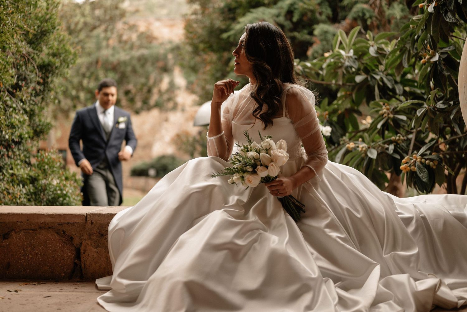 A bride in a flowing white gown sits on stone steps holding a bouquet while a suited figure approaches in the background.