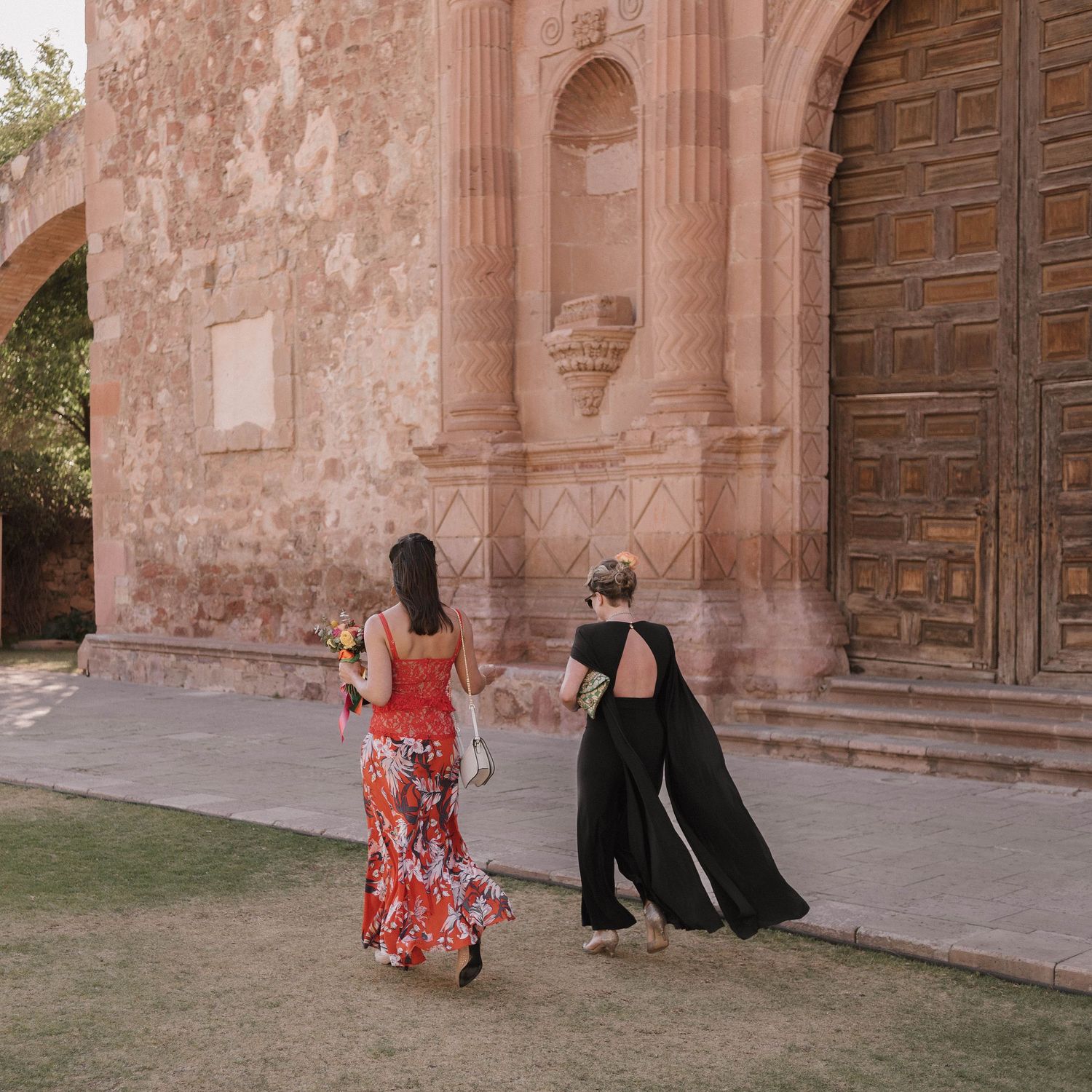 Two women in flowy dresses walk near a historic mission building with pink stone walls.