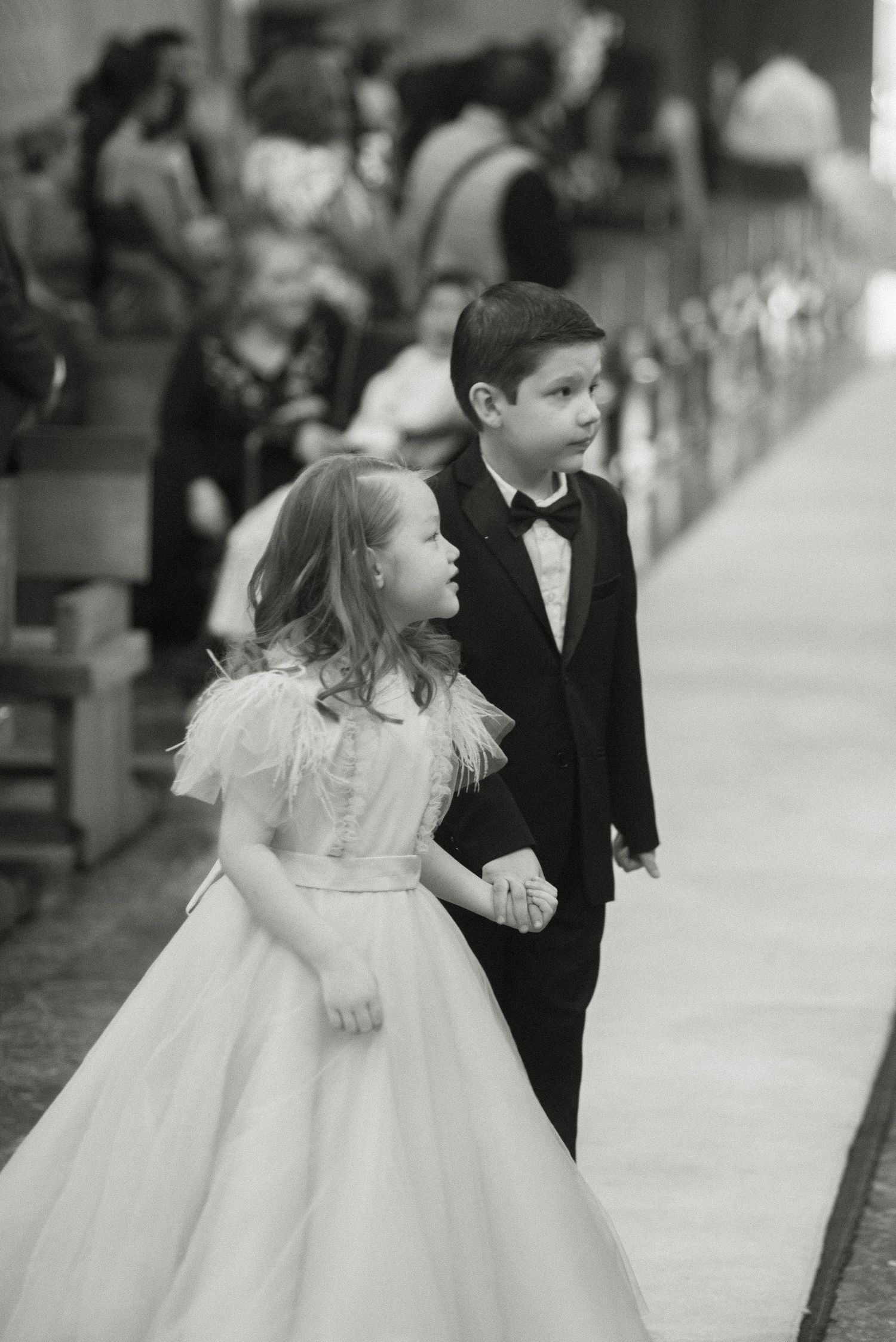 A young boy in a tuxedo and girl in a white dress walk together down an aisle at a formal event.