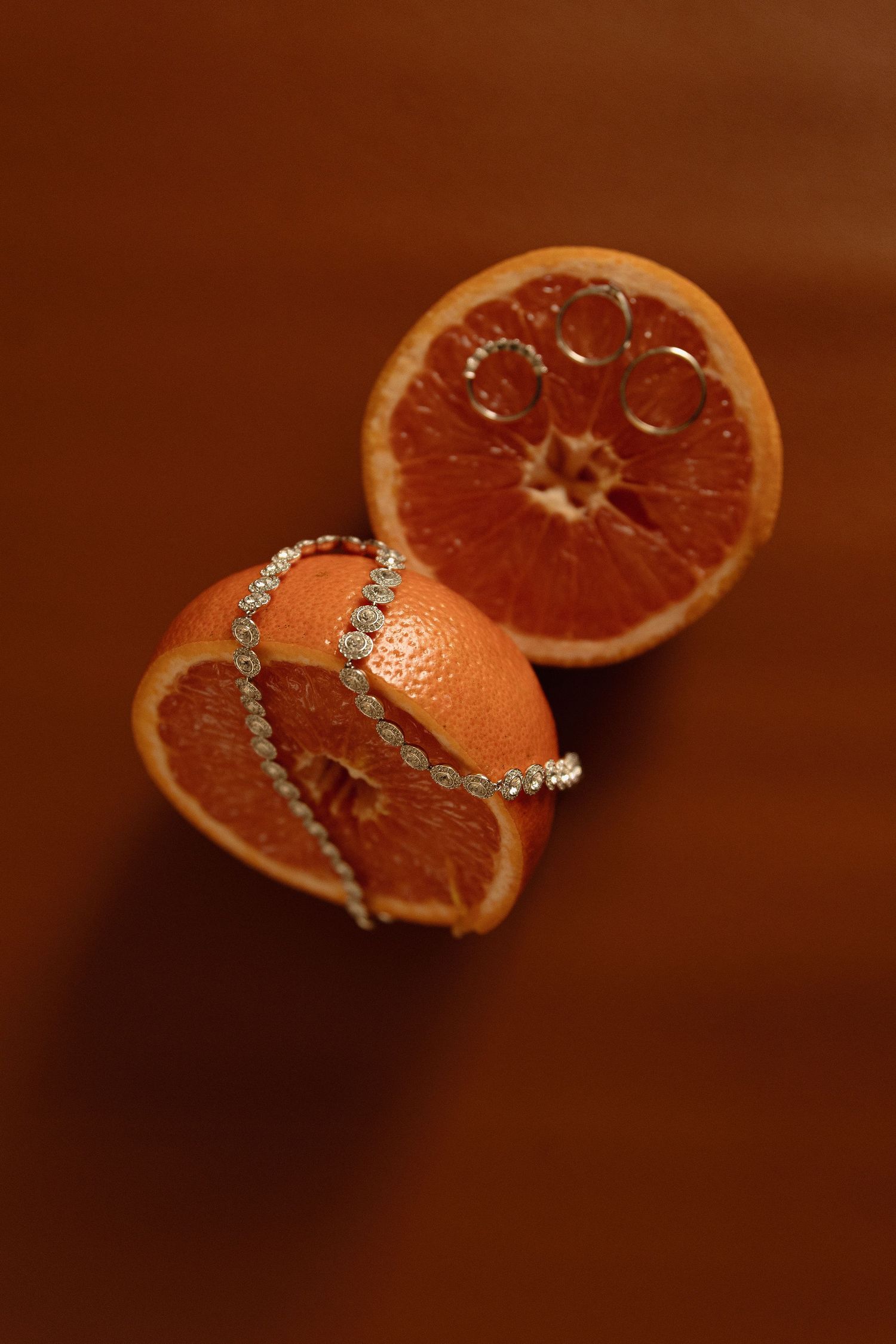 Two halves of a blood orange adorned with diamond jewelry rings on a rich brown background.