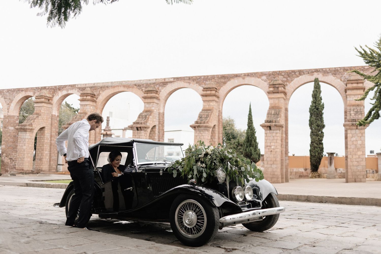 A vintage black car parked beneath stone arches with cypress trees in the background at a historic location.
