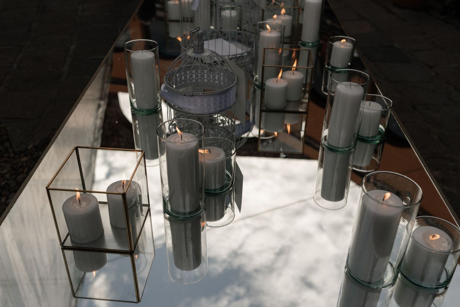 Assorted silver glass vases and candle holders arranged on reflective white marble surface with dramatic lighting.
