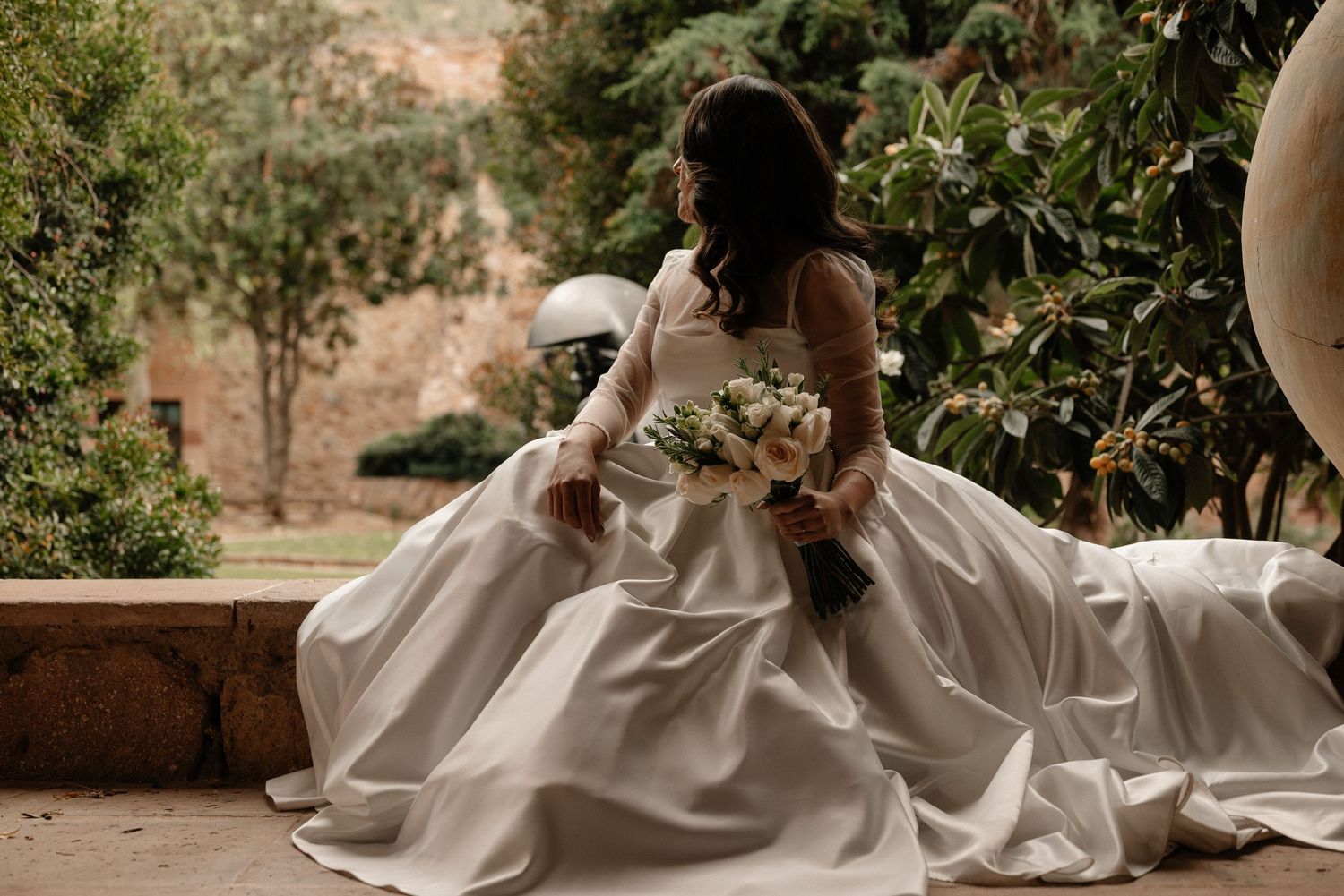 Bride in a flowing white wedding dress sits on stone steps surrounded by lush greenery in an outdoor garden setting.
