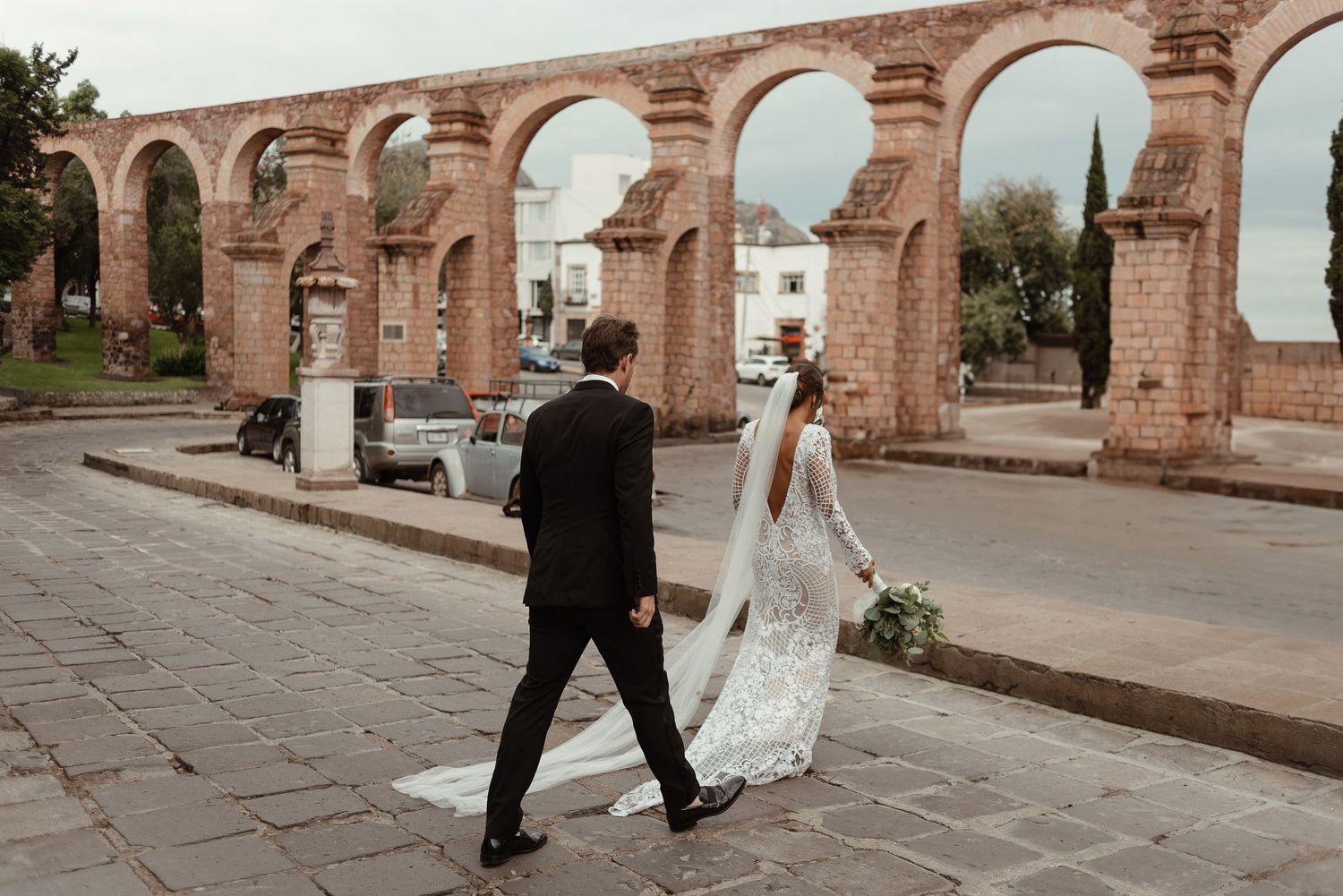 Wedding couple walks under elegant brick archways in a historic courtyard.