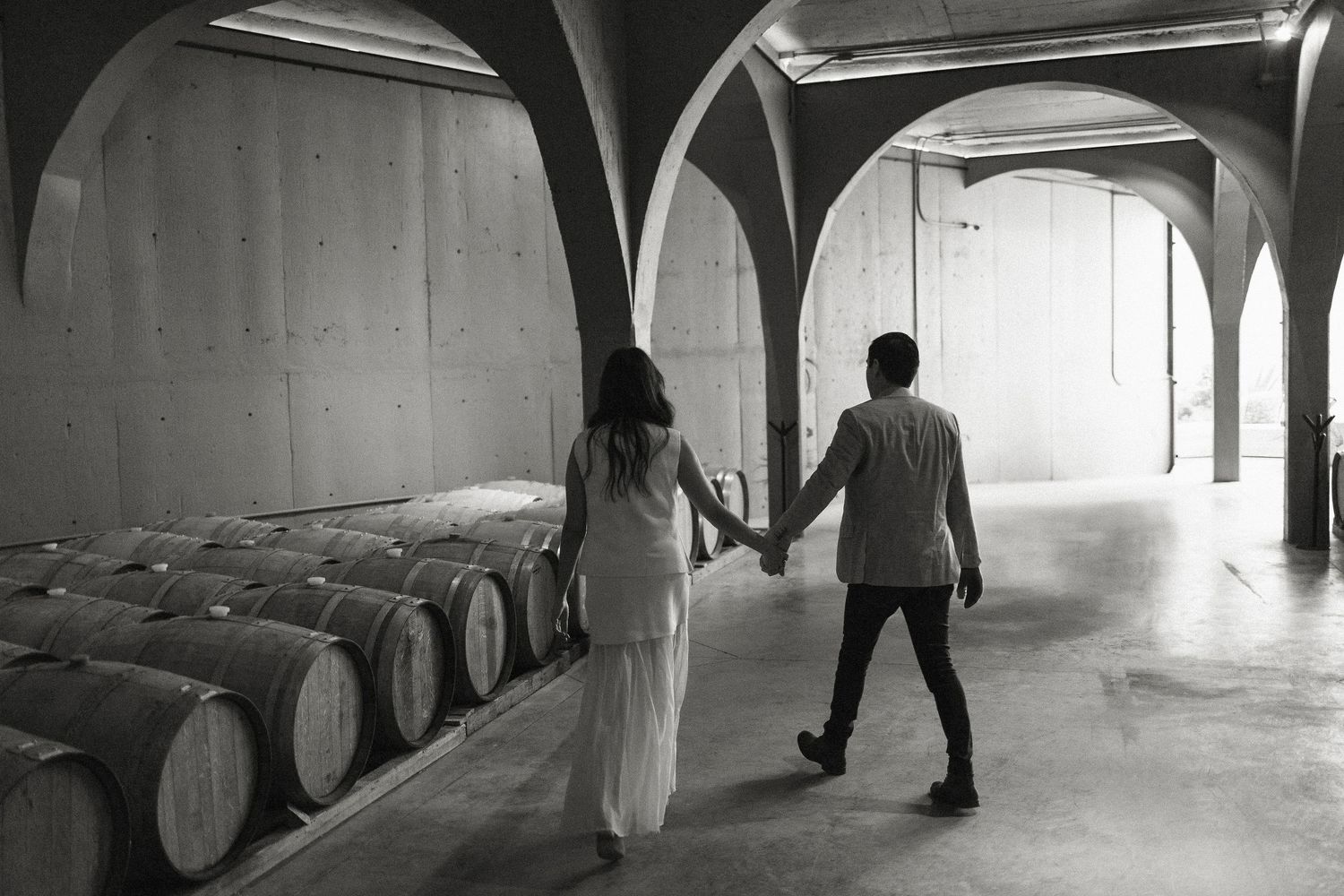 Black and white photograph of people walking through a dimly lit wine cellar with rows of wooden barrels.