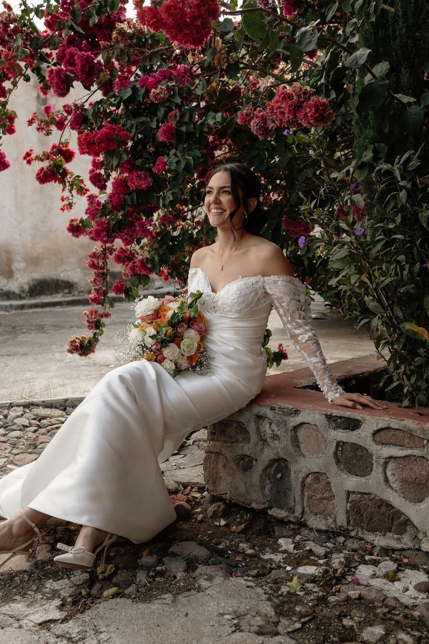 Elegant bridal portrait with flowing white dress against vibrant red bougainvillea flowers and stone wall.