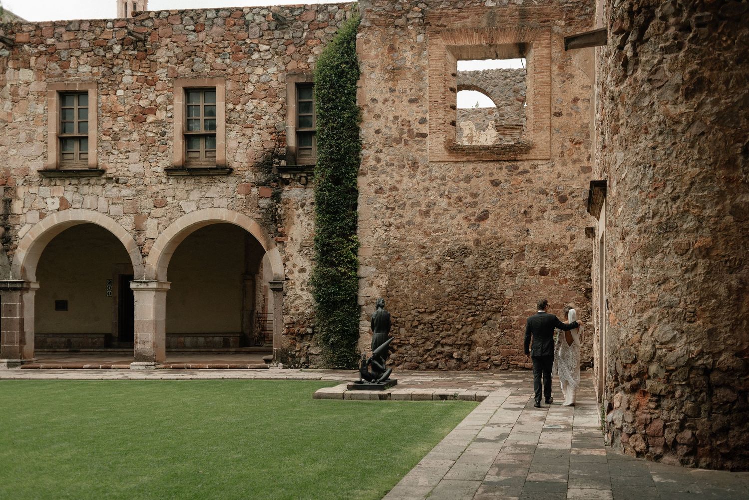 Two people stand near stone archways and ivy-covered walls in a historic courtyard with green grass.