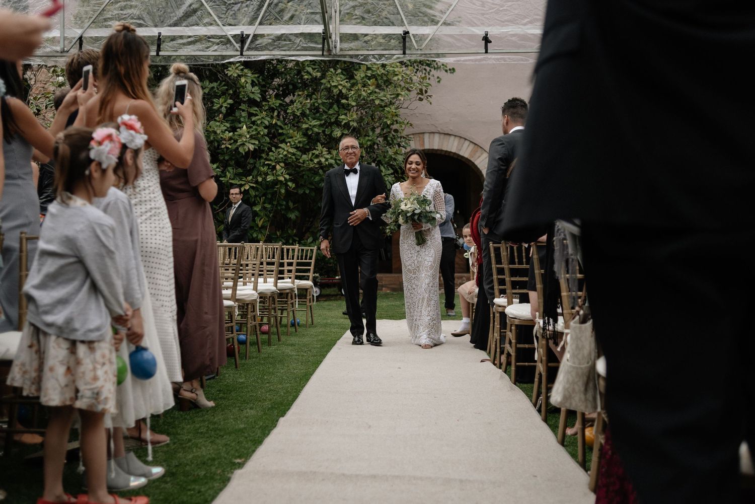A wedding ceremony takes place outdoors with guests lining a white aisle runner as a couple walks down together.