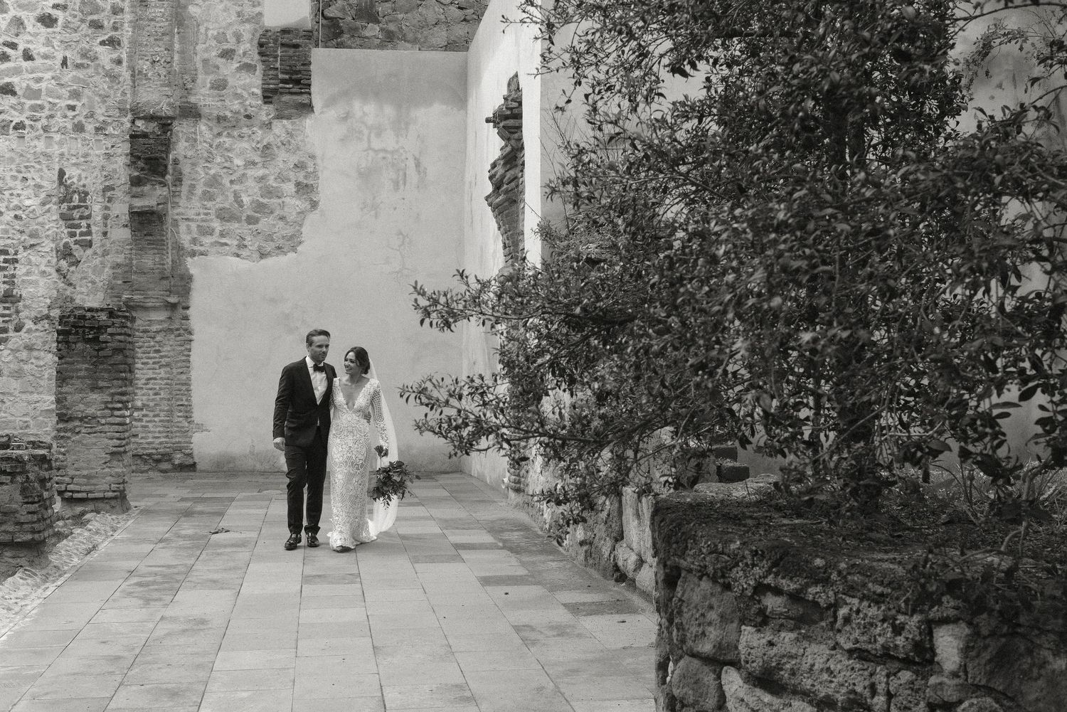 A couple walks along a stone pathway beside old brick ruins and trees in black and white.