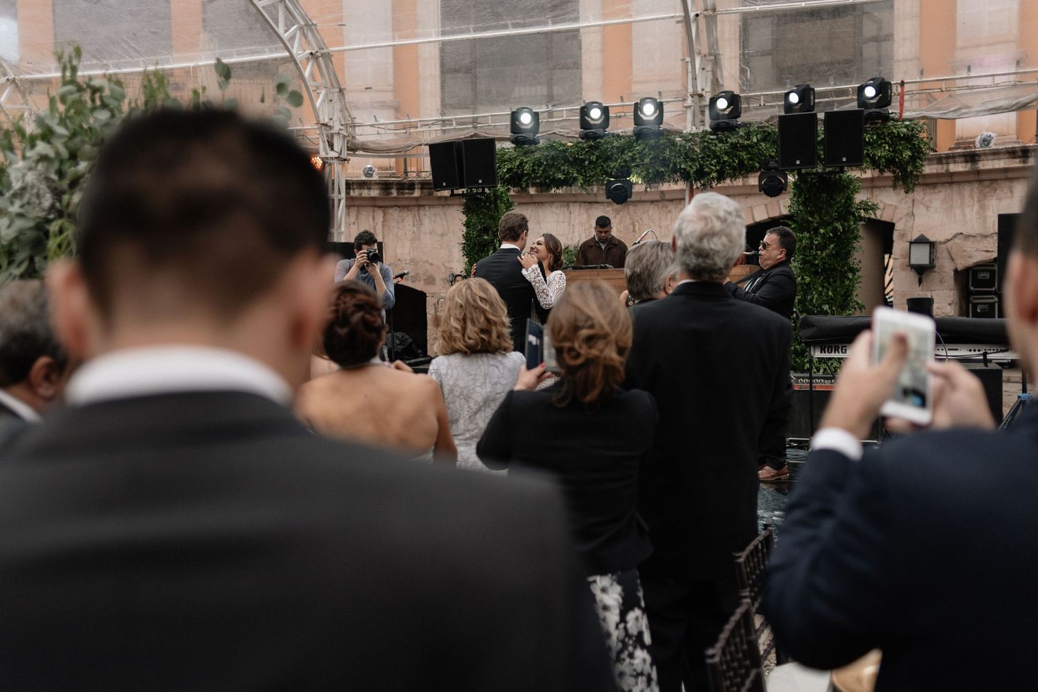Wedding guests take photos of a ceremony in a courtyard decorated with greenery and string lights.