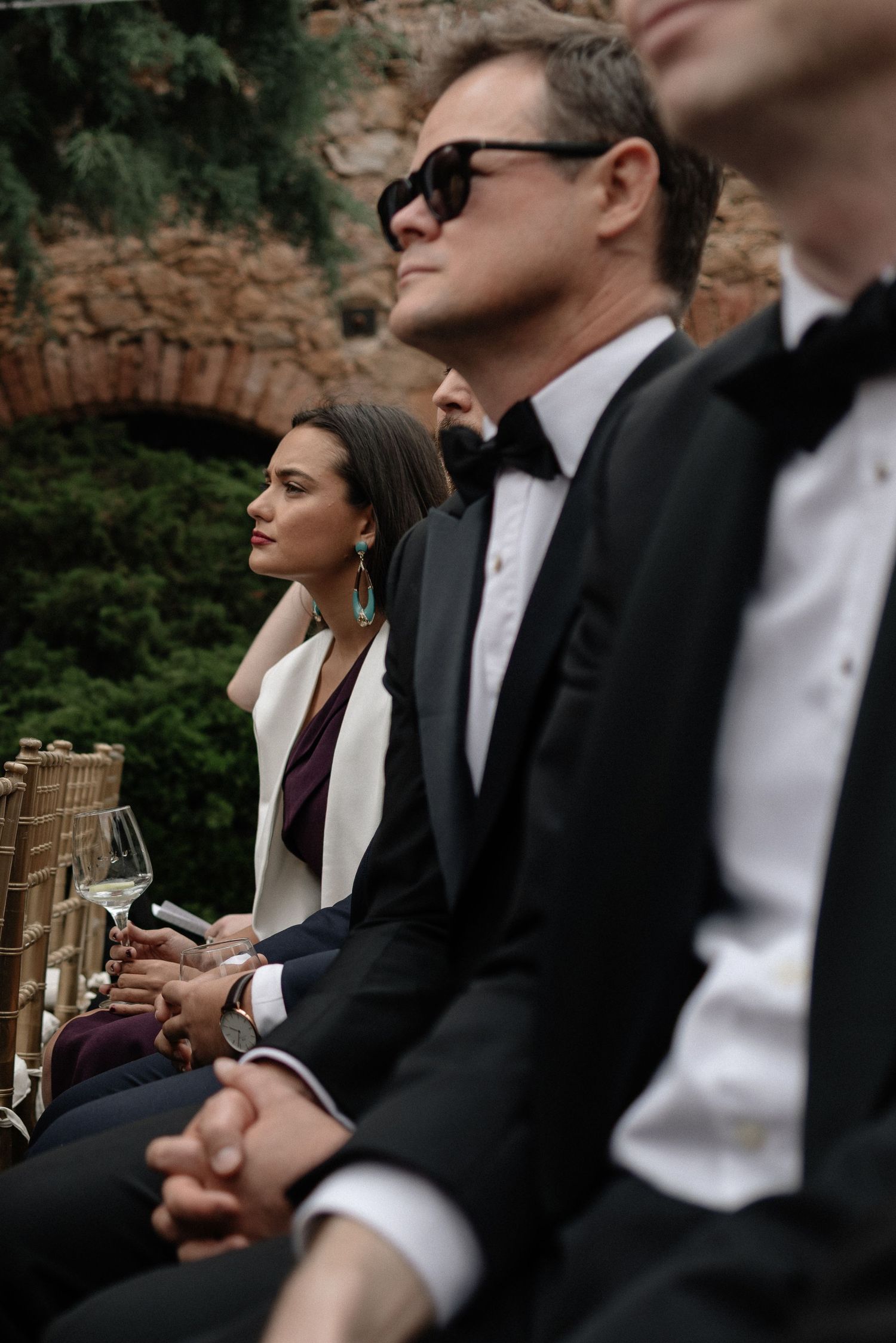 Elegantly dressed guests in formal attire sit during an outdoor event with wine glasses in hand.
