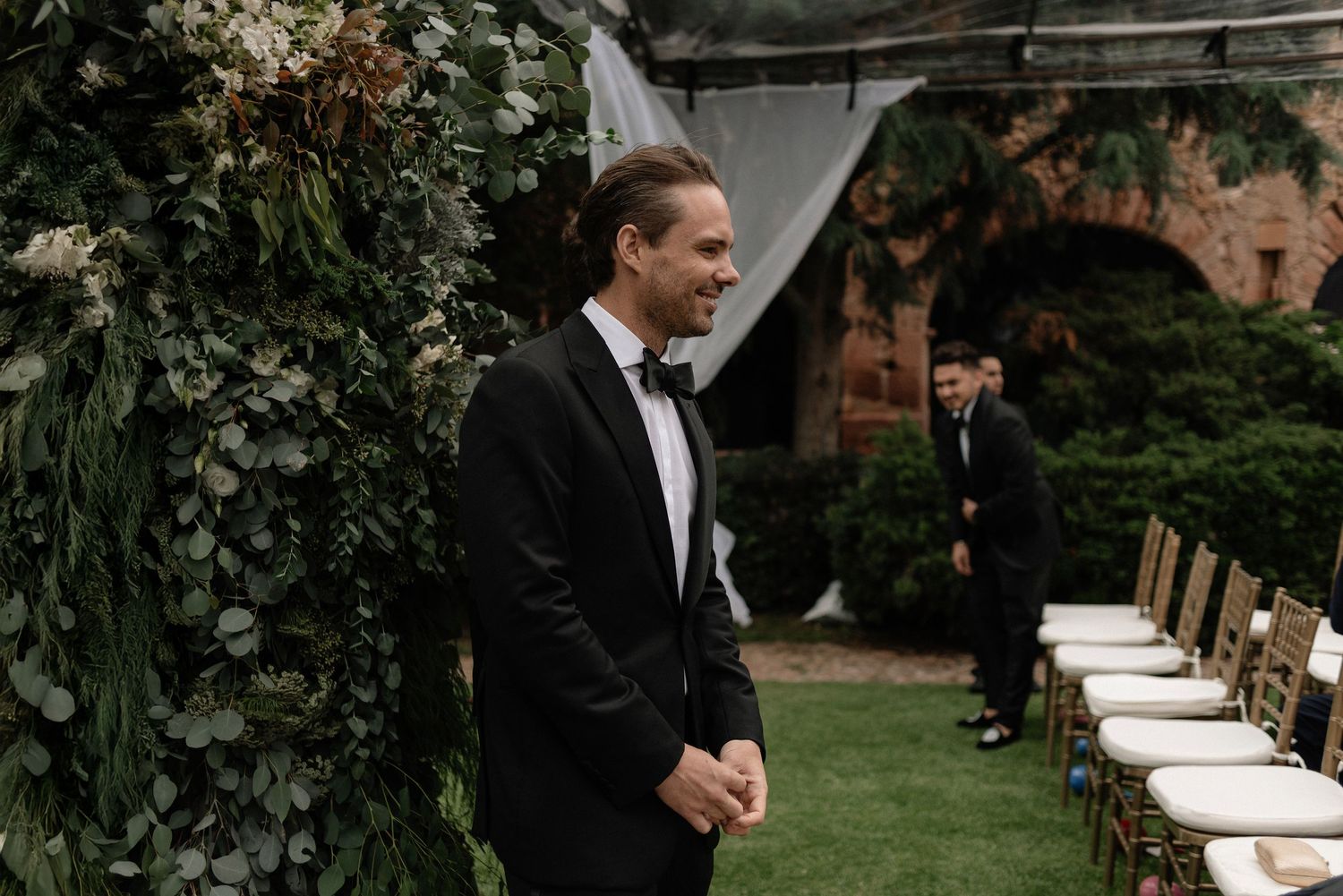 A groom in a black tuxedo and bowtie stands by a floral arch at an outdoor garden wedding ceremony setup.
