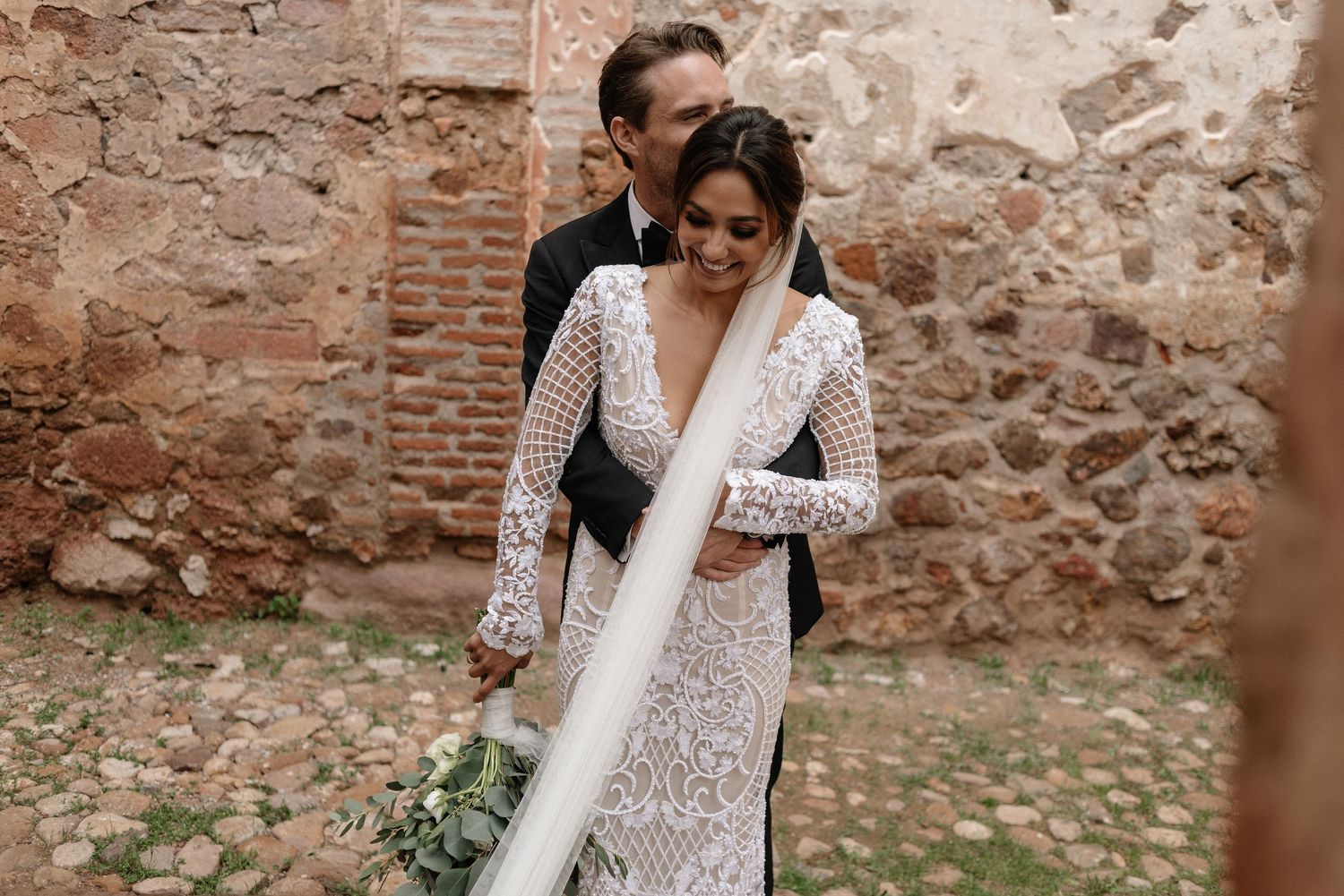A couple shares an intimate embrace at their wedding, with the bride wearing an elegant long-sleeved lace gown against stone walls.