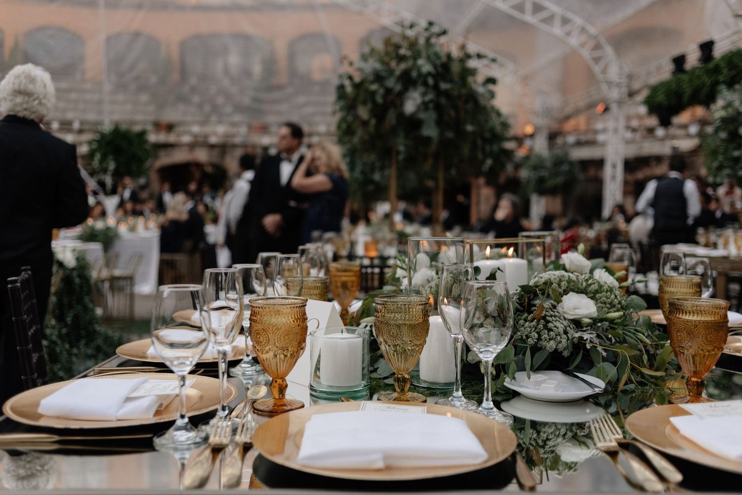 Elegant wedding reception table set with amber glassware, white plates, and lush greenery centerpieces in an ornate venue.
