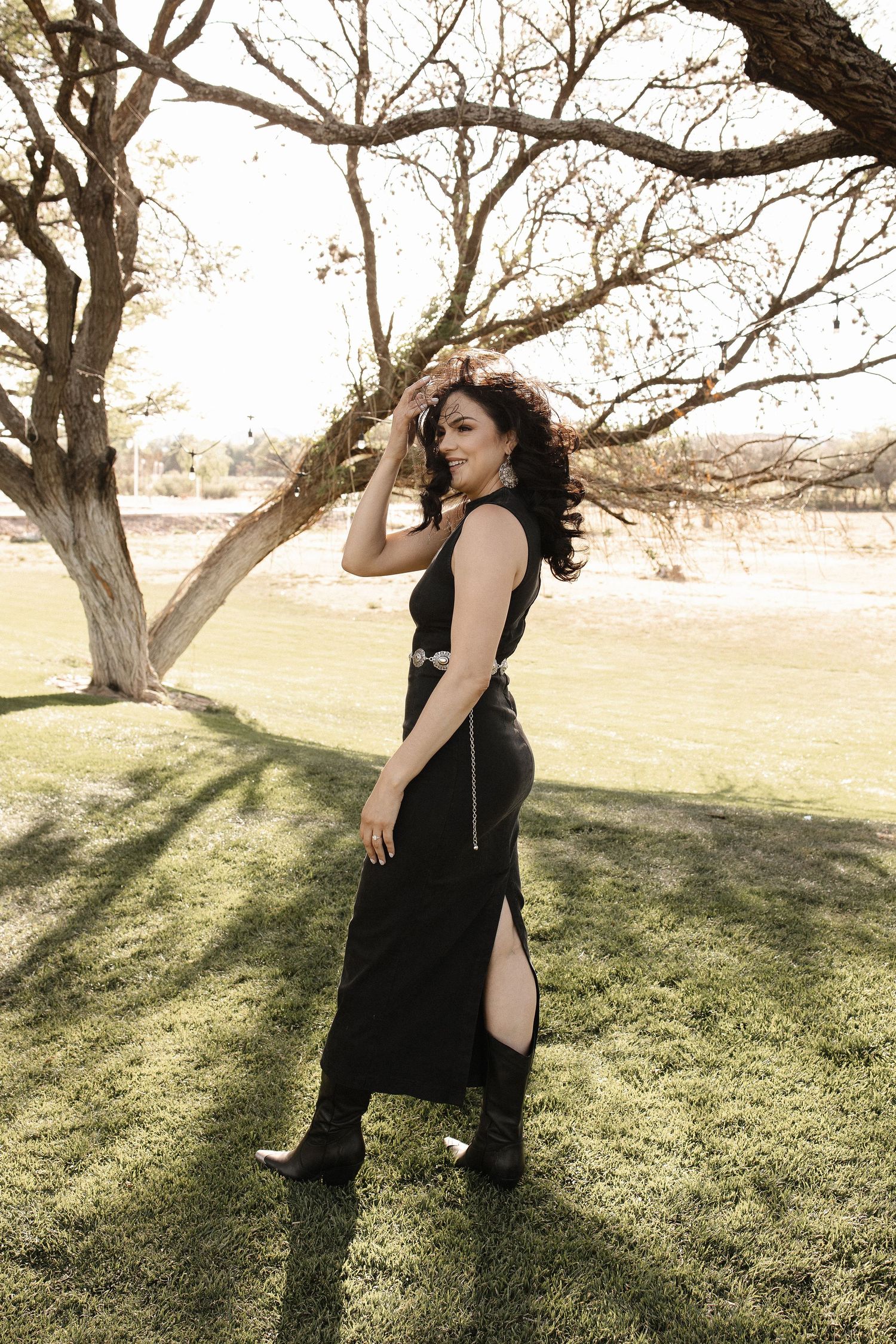 A figure in a black sleeveless dress poses outdoors near a bare tree on a grassy lawn in soft sunlight.