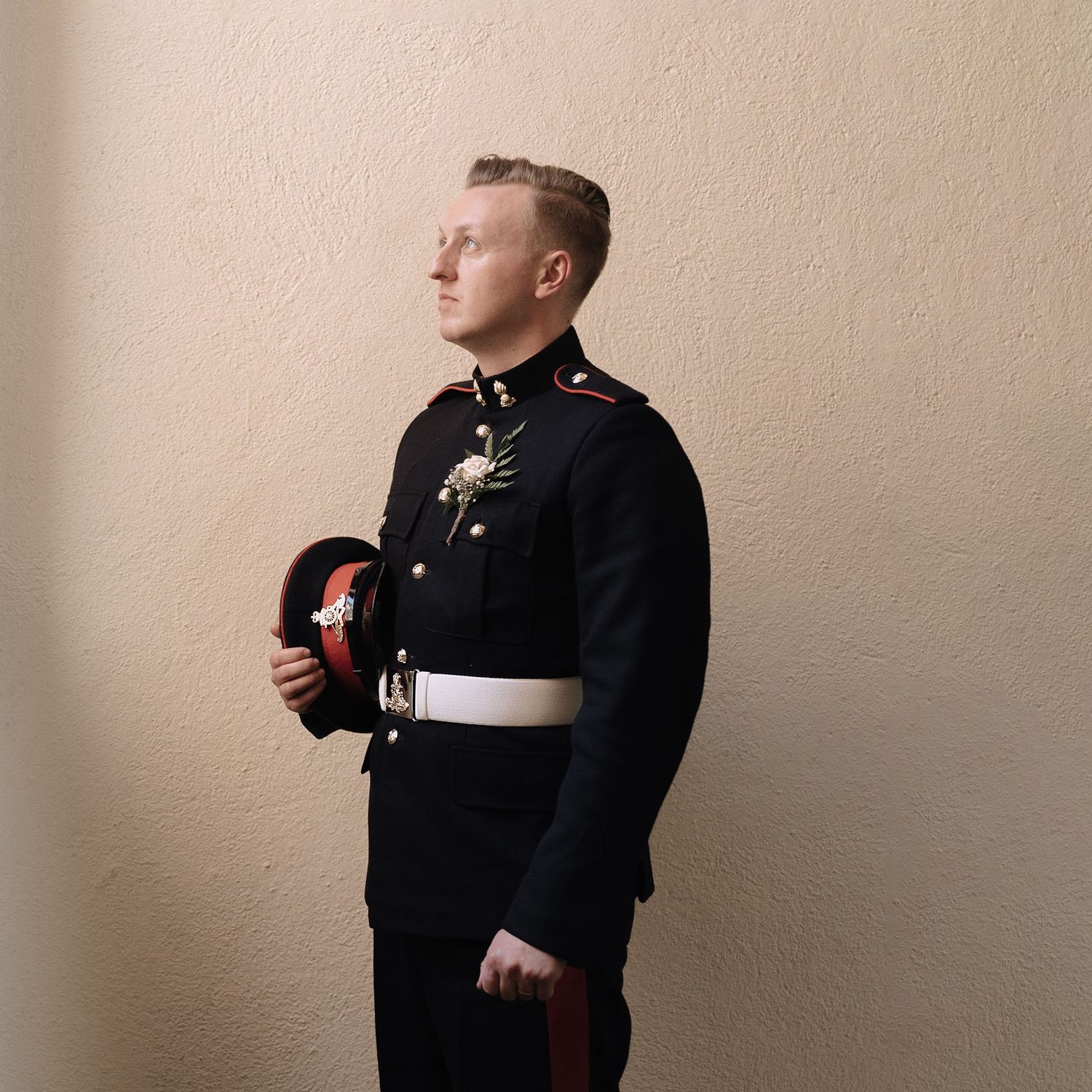 Stylish groomsman in black suit adjusts boutonniere against neutral wall.