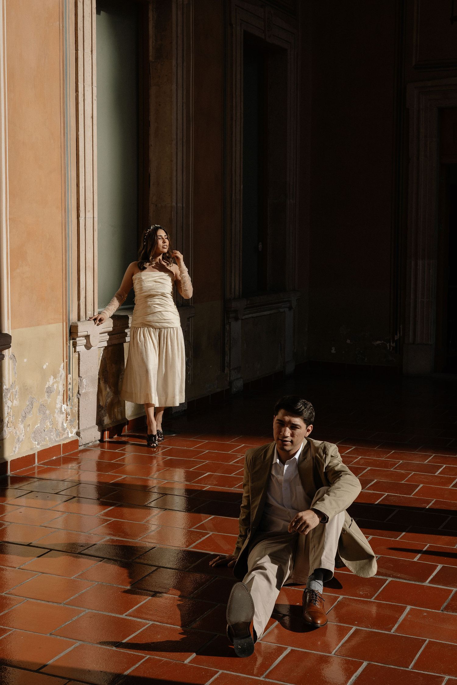 Two people in vintage style clothing pose dramatically in a sunlit hallway with terracotta floors and dark walls.