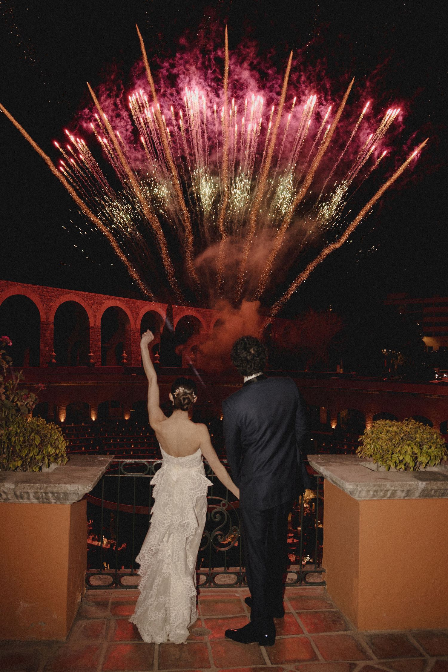 Couple watching spectacular pink and gold fireworks display over an arched bridge at night during wedding celebration.