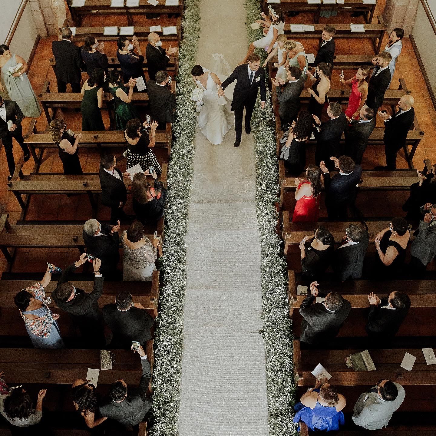 Overhead view of wedding ceremony in church with white aisle runner and wooden pews filled with guests.