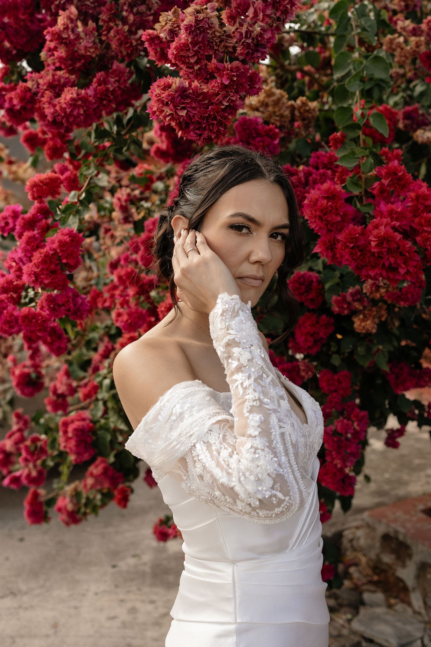 A figure in a white lace dress poses against vibrant red bougainvillea flowers in a romantic outdoor setting.