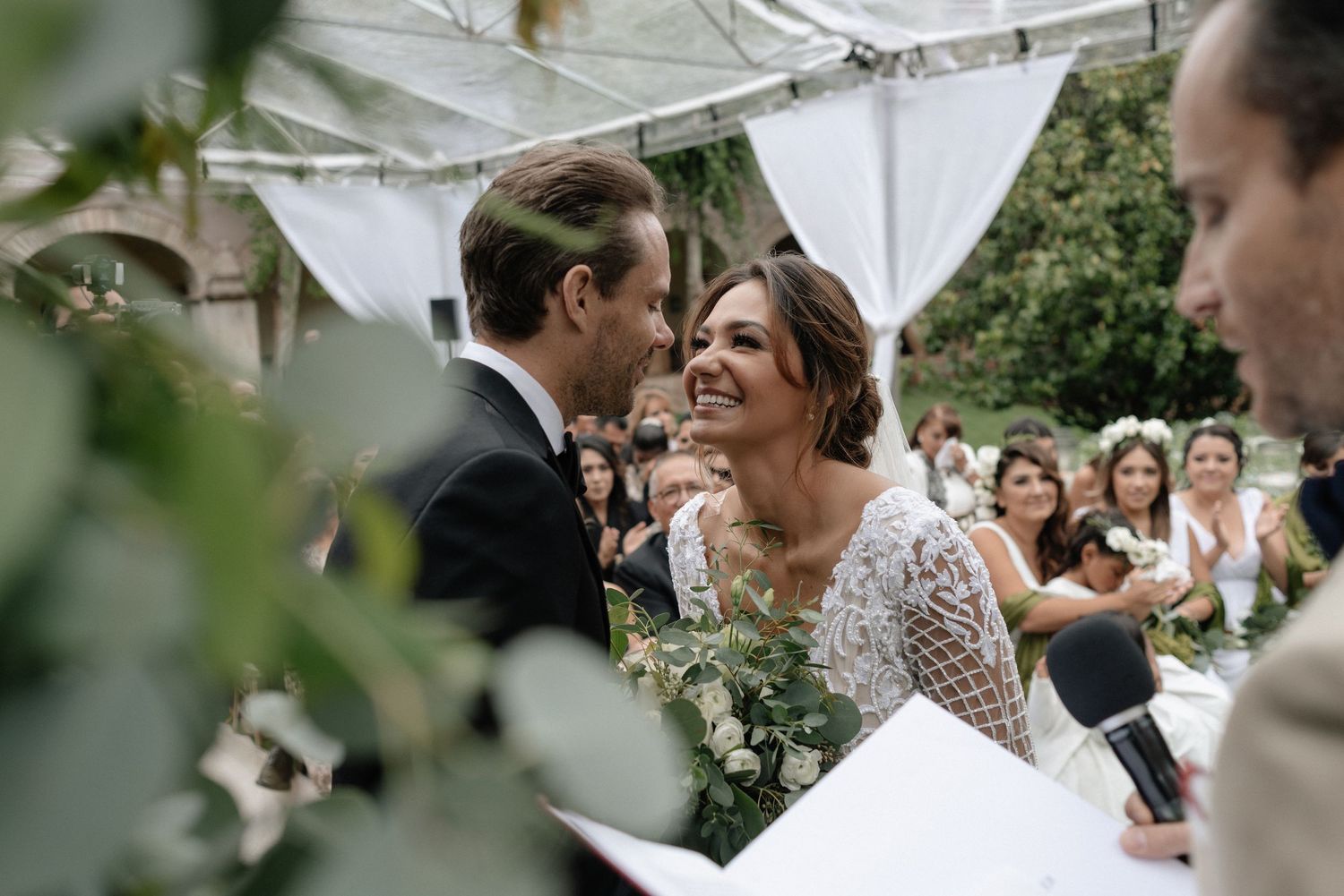 Couple shares joyful moment during outdoor wedding ceremony under white tent canopy with guests looking on.