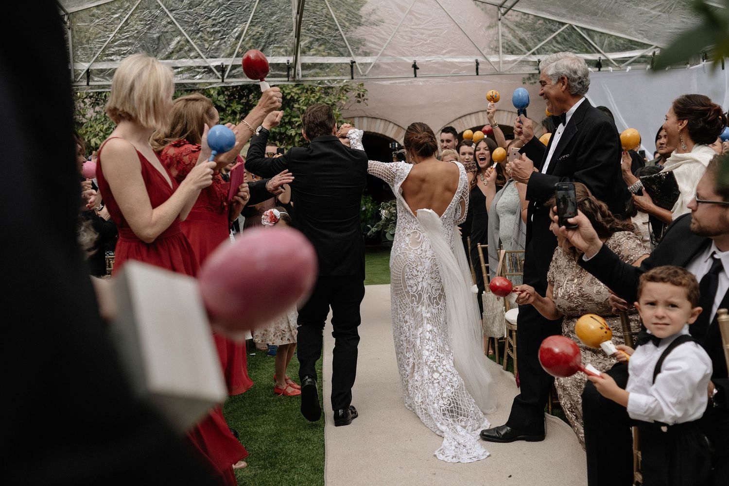 Wedding guests blow bubbles as newlyweds walk down the aisle under a tent during an outdoor ceremony.
