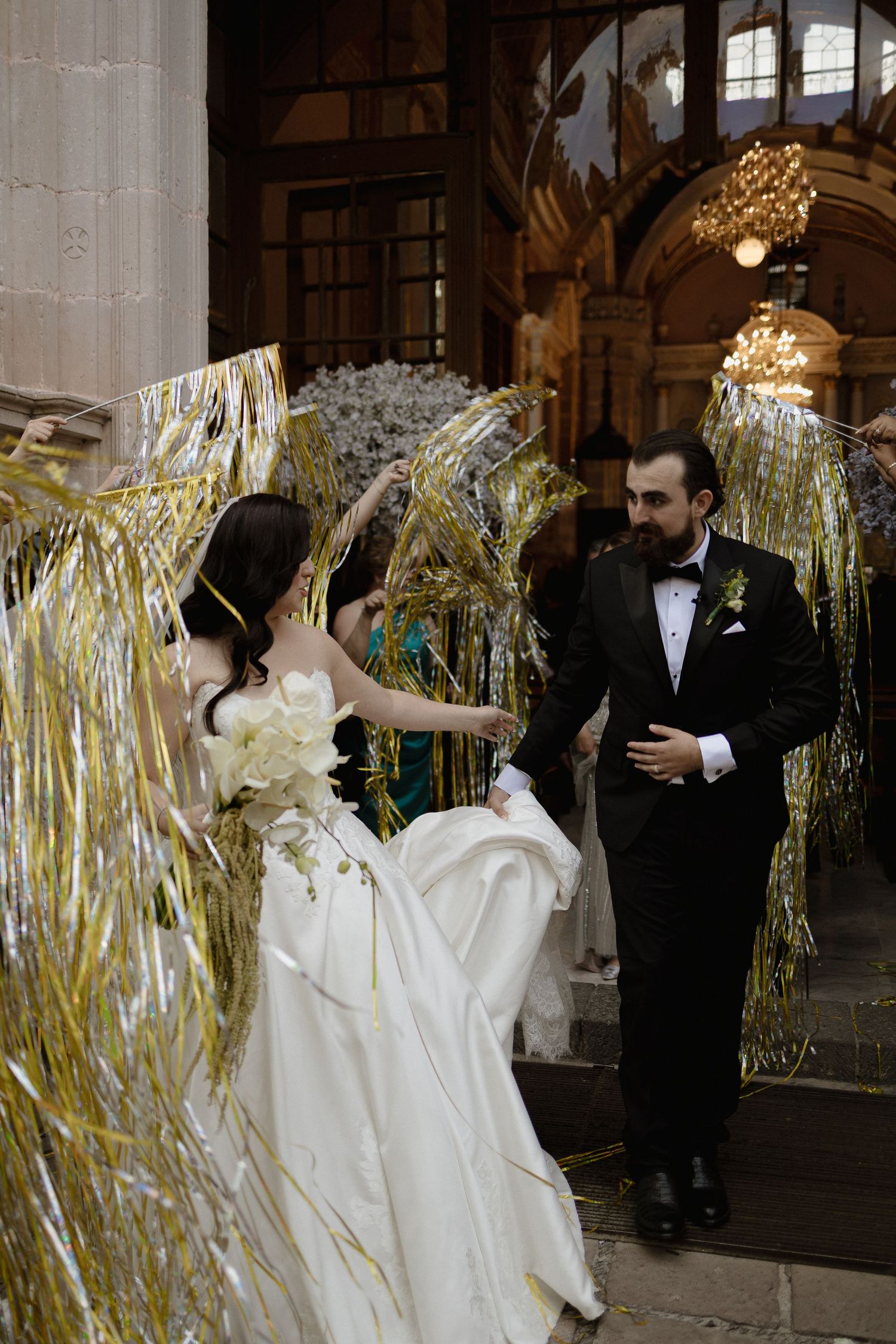 Bride and groom exit church through gold tinsel curtain, bride in white gown, groom in black tuxedo.