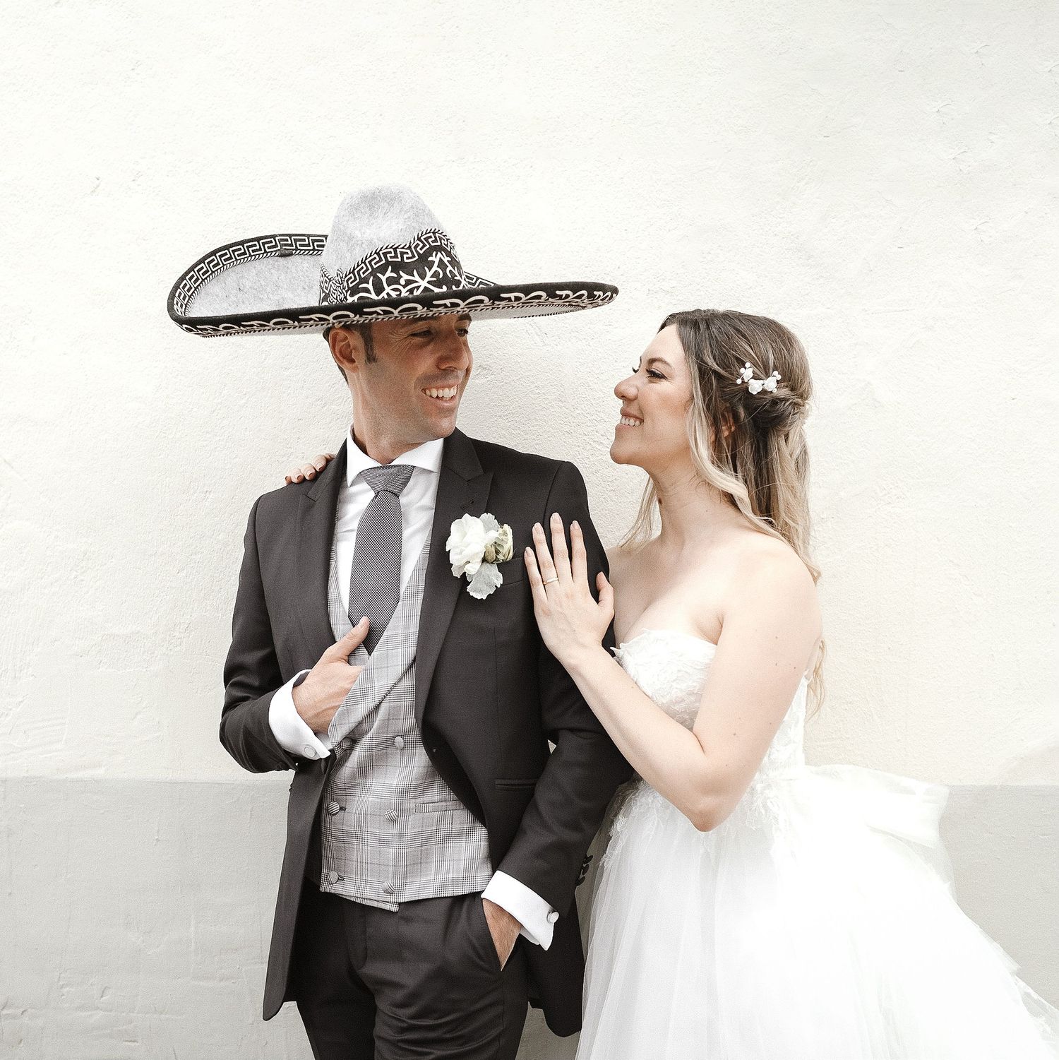 Wedding couple in formal attire with groom wearing decorative Mexican sombrero against white wall.