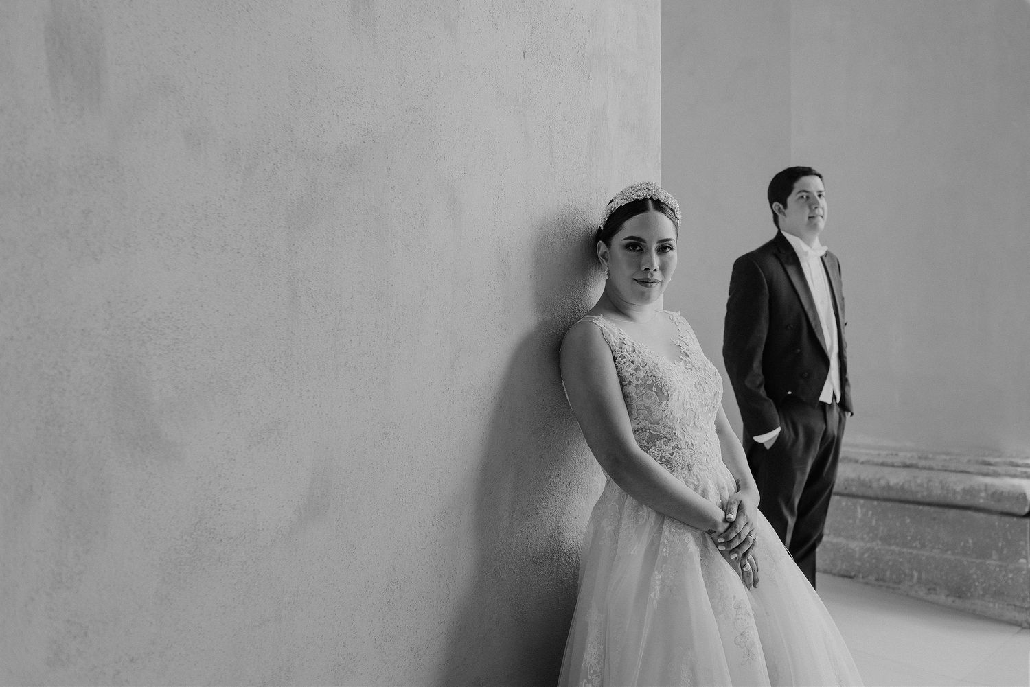 Black and white portrait of couple in wedding attire standing against a minimalist wall.