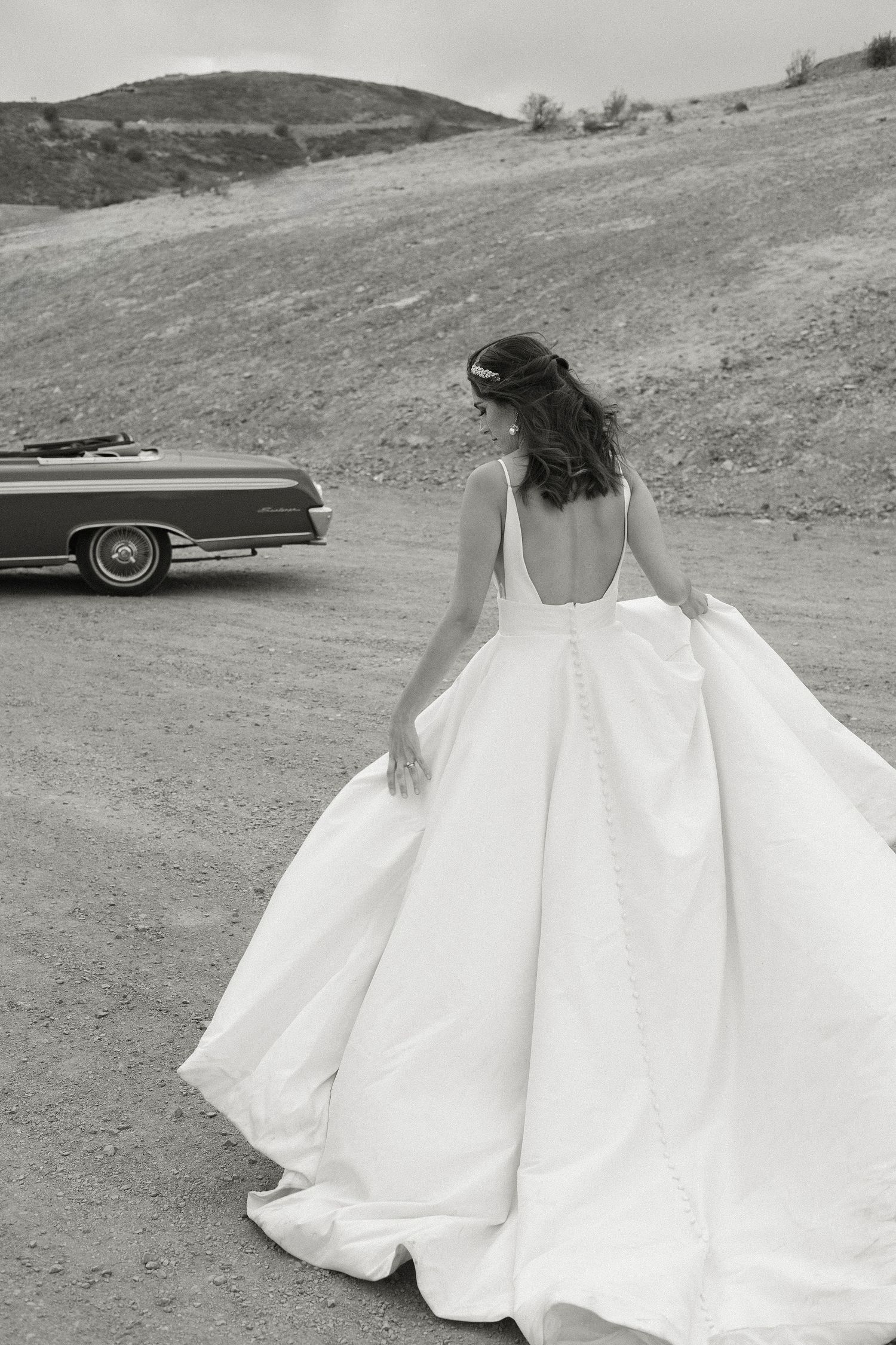 A bride in a flowing white ball gown poses near a vintage car on a dirt road against rolling hills.